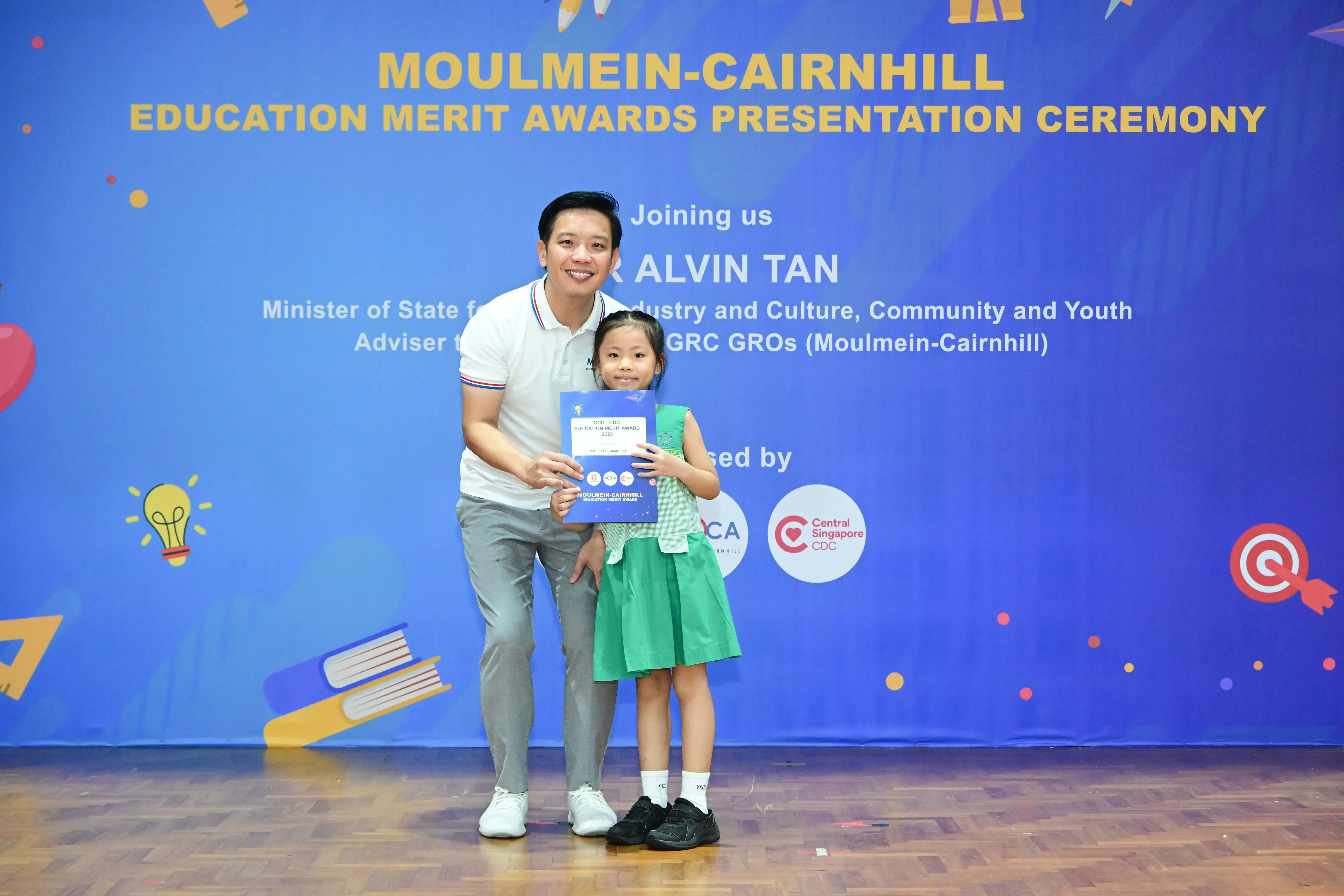 A man and a young girl in green hold a certificate at the Moulmein-Cairnhill Education Merit Awards Ceremony.