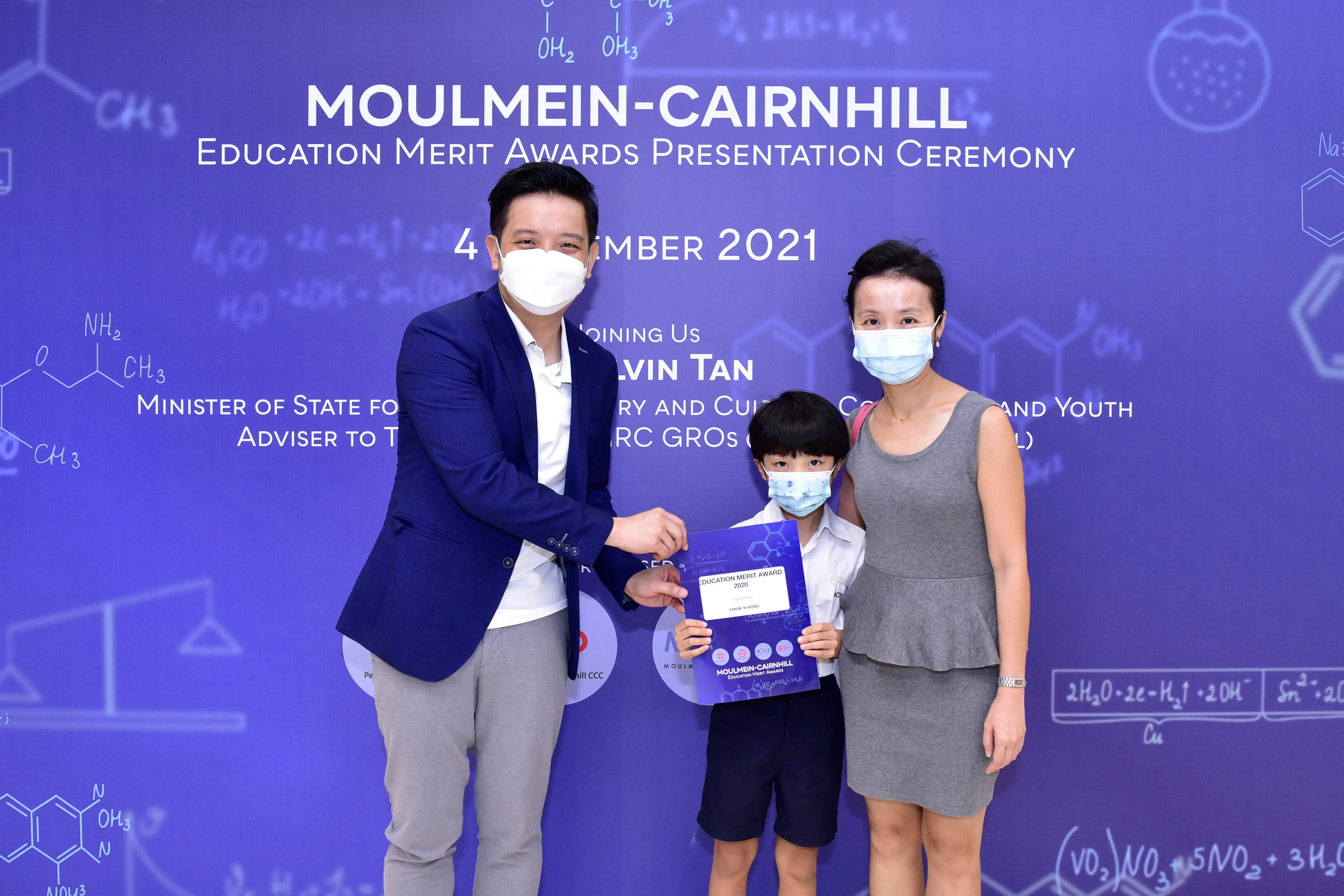 Three masked people pose at the Moulmein-Cairnhill Education Merit Awards 2021, with a child holding a certificate.