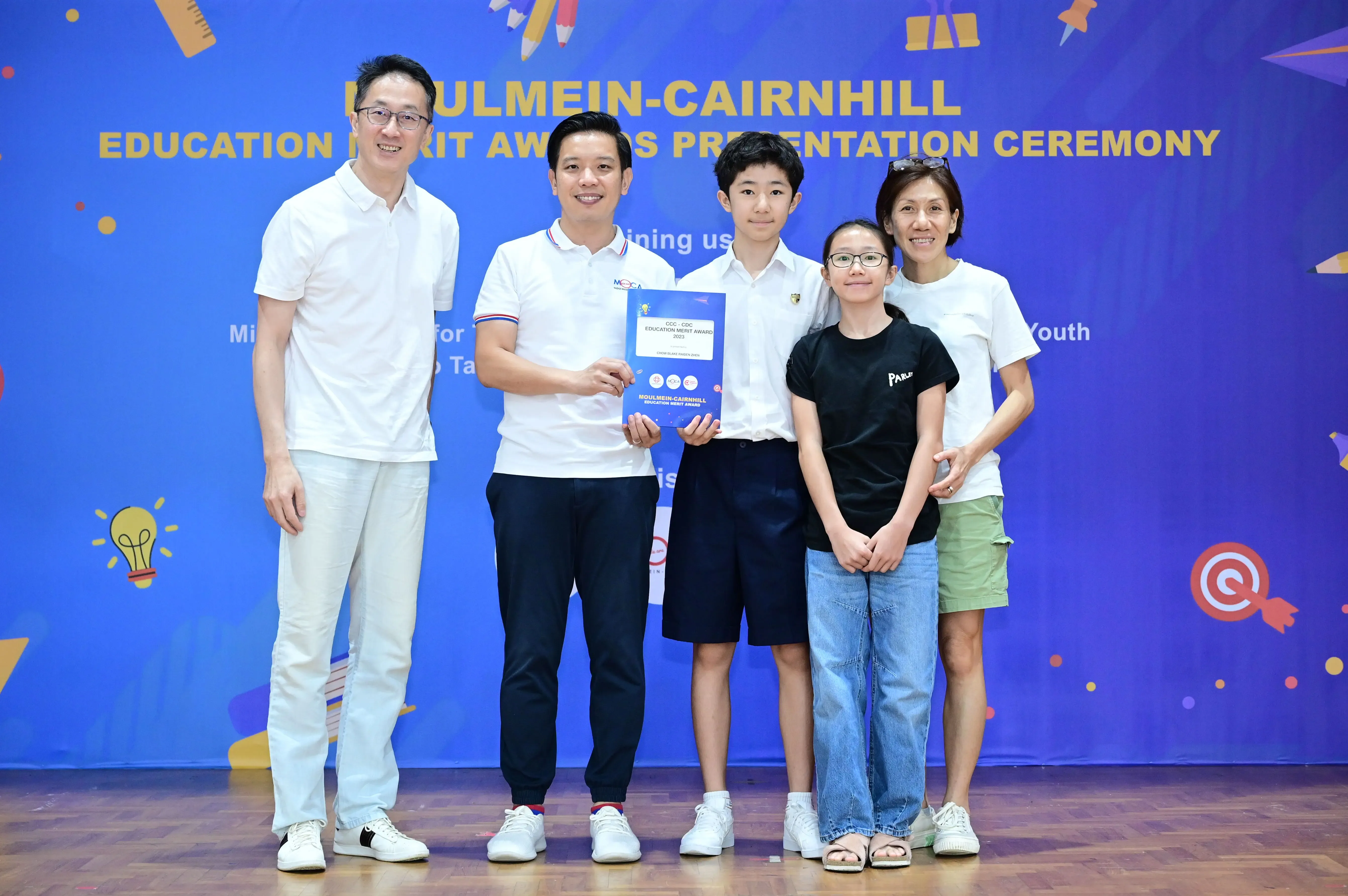 Five people pose on stage at the Moulmein-Cairnhill Education Merit Awards Presentation Ceremony, holding a certificate.