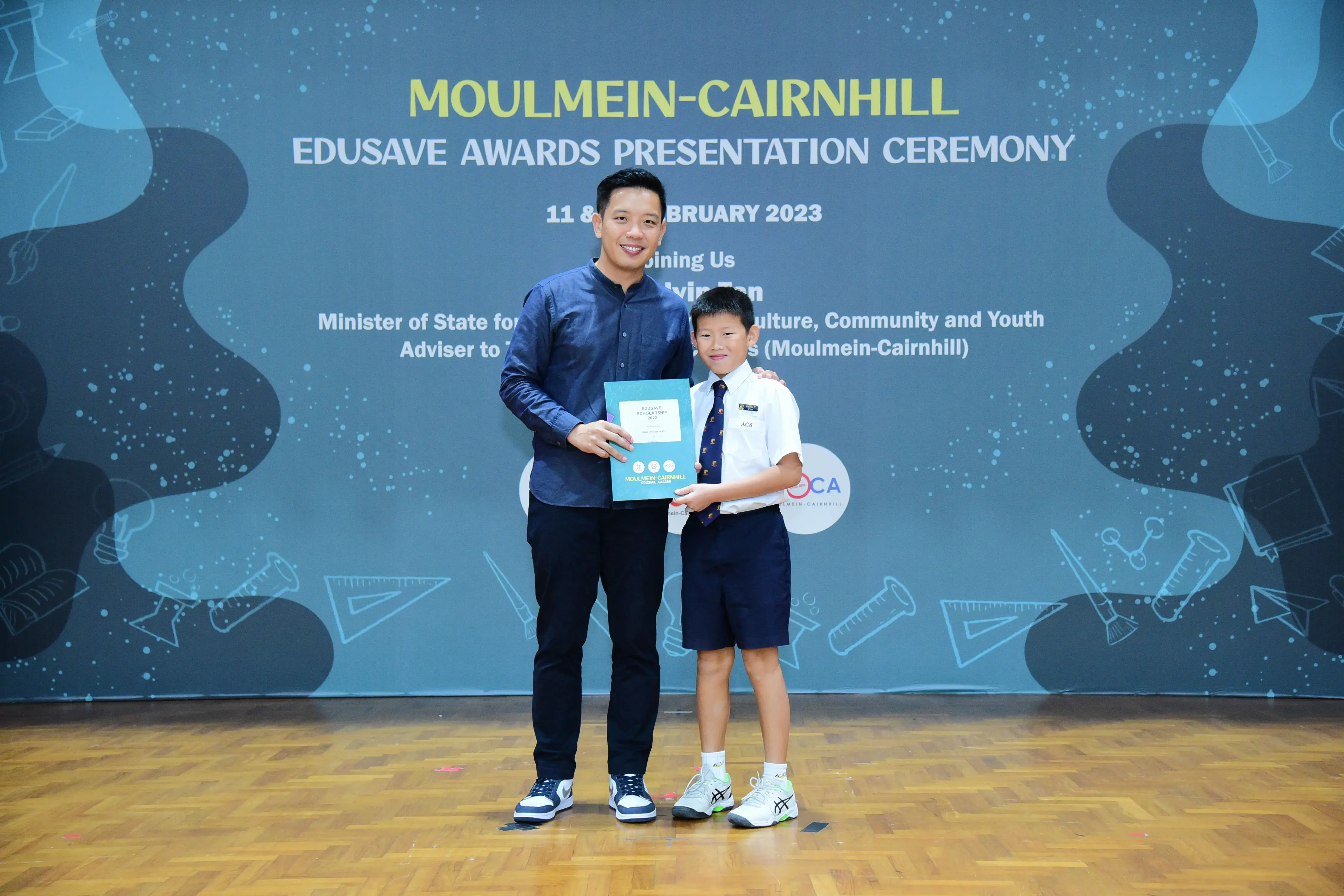 A man and a young boy in school uniform hold a certificate at the Moulmein-Cairnhill Edusave Awards Ceremony 2023.