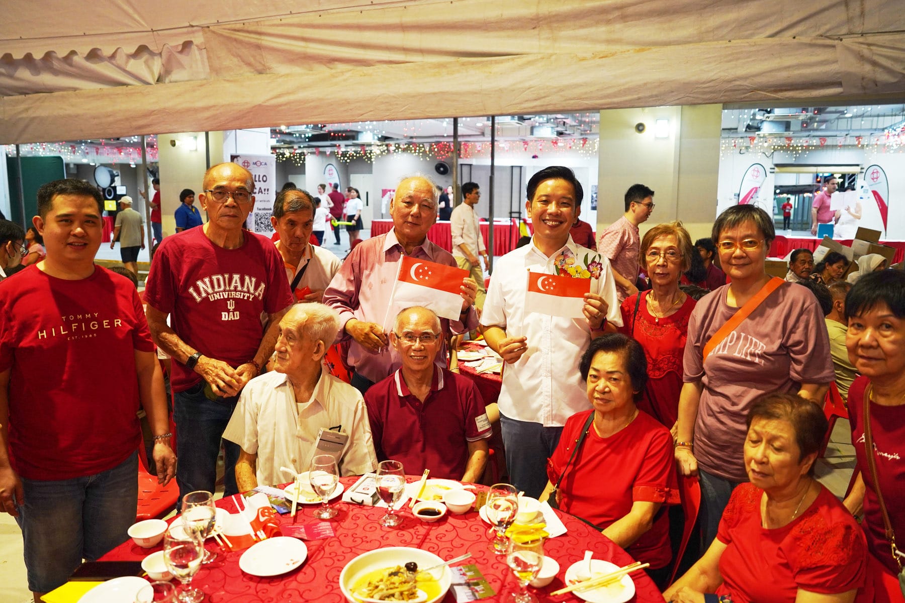 A group of people in red clothing pose around a dining table, with two men holding small Singapore flags.