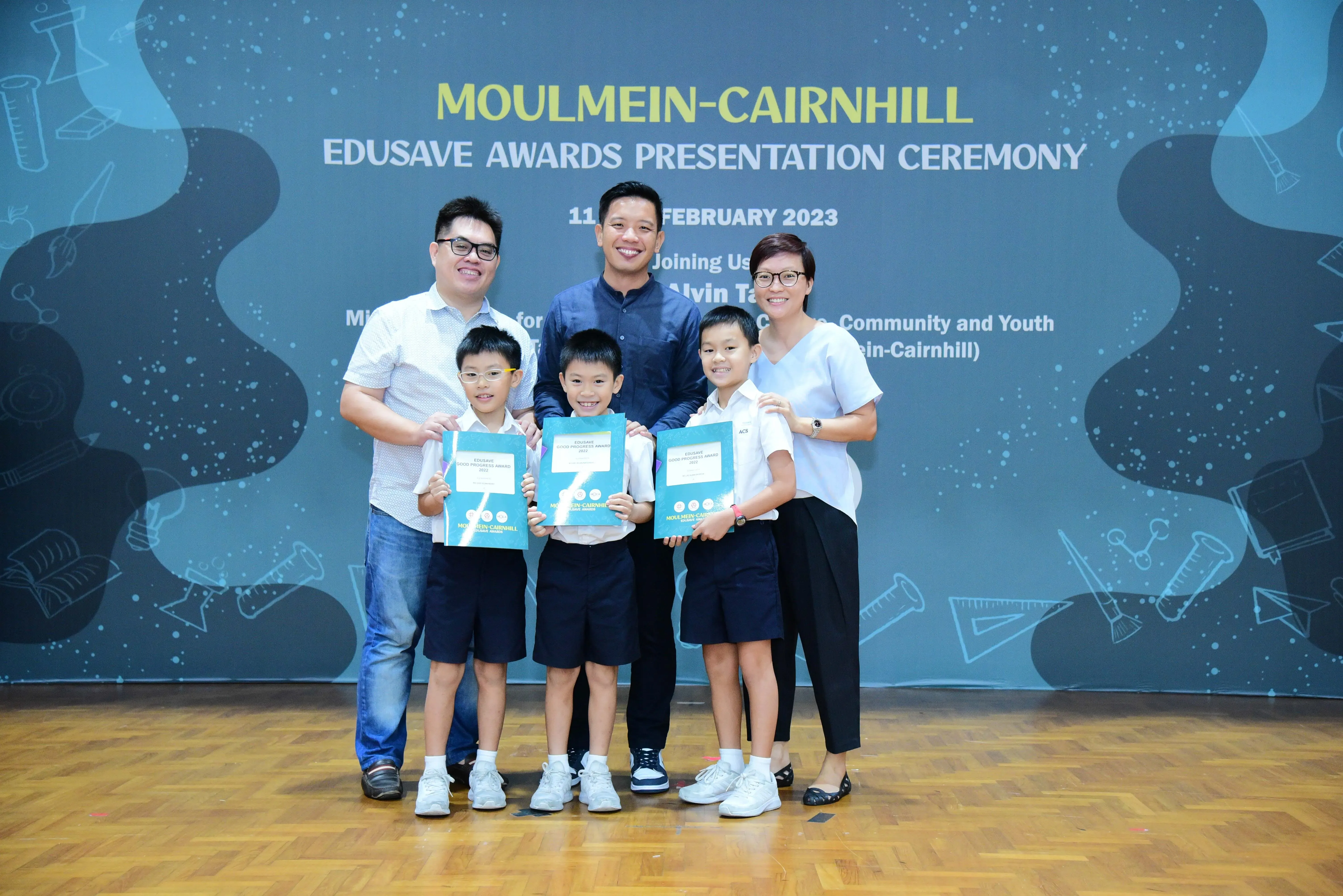 Three boys holding award booklets pose with two adults at the Moulmein-Cairnhill Edusave Awards Ceremony 2023.