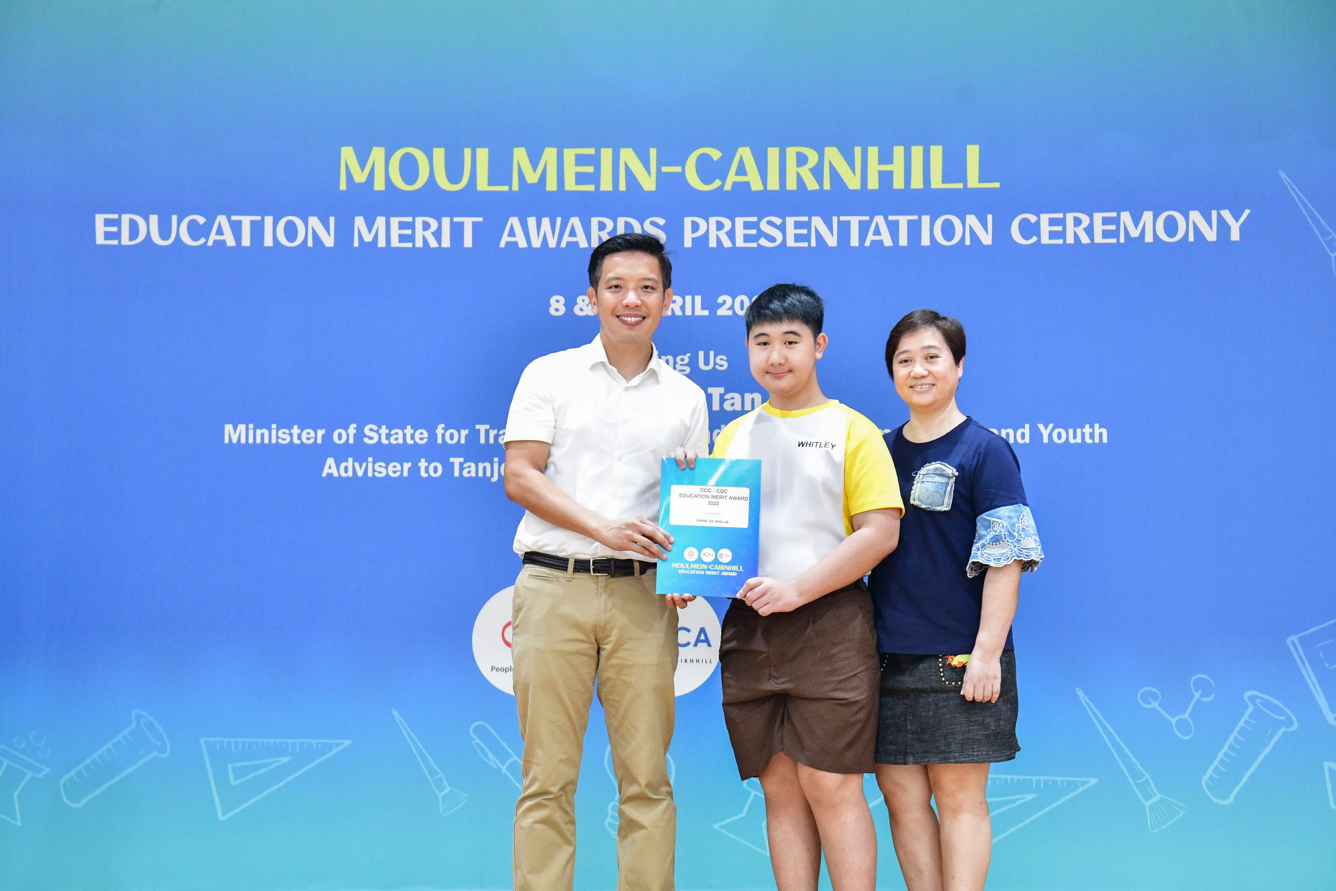 Three people pose at the Moulmein-Cairnhill Education Merit Awards Presentation Ceremony, with a student holding an award.