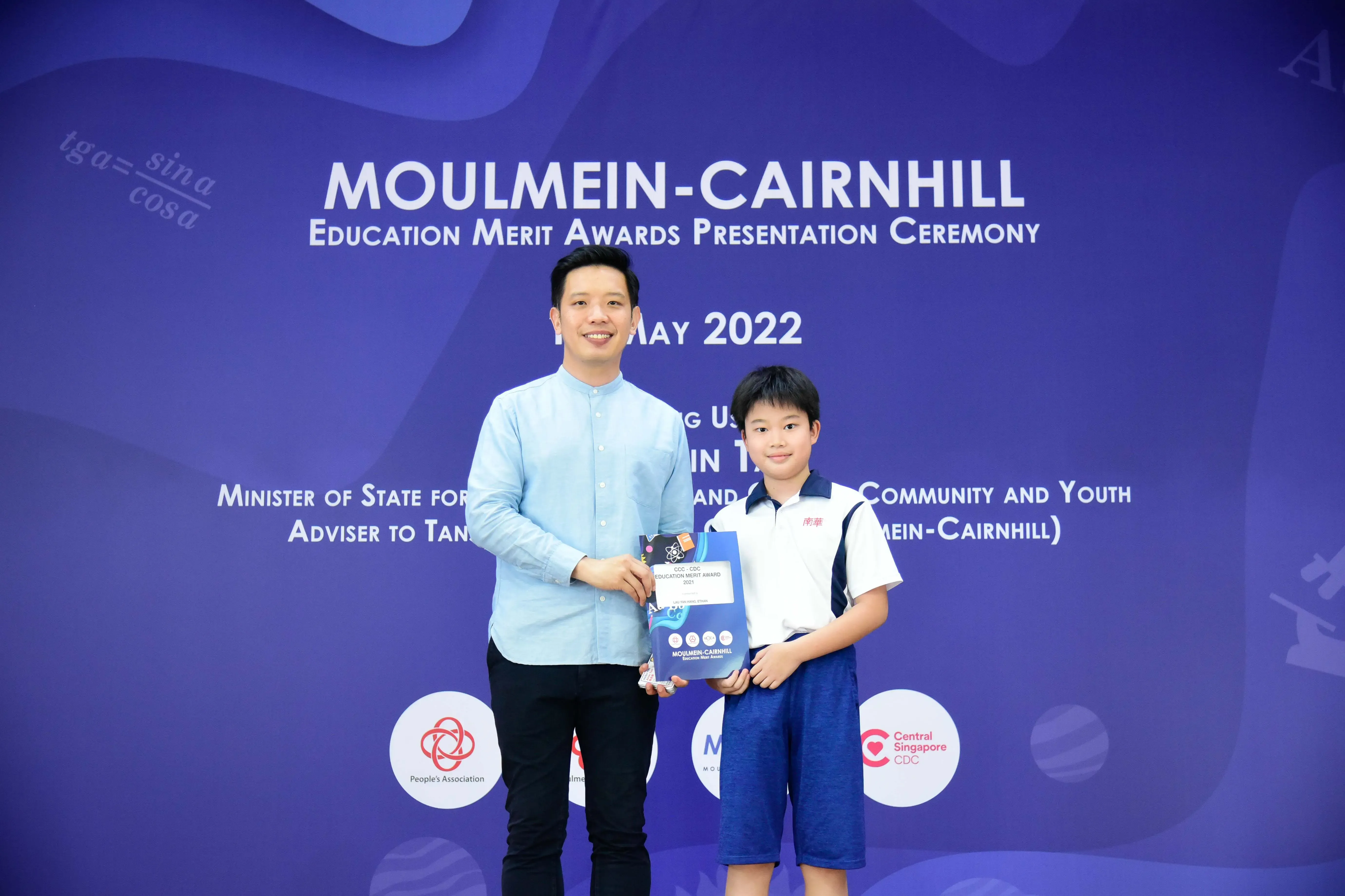 A man and a young boy hold an award packet at the Moulmein-Cairnhill Education Merit Awards Ceremony, May 2022.