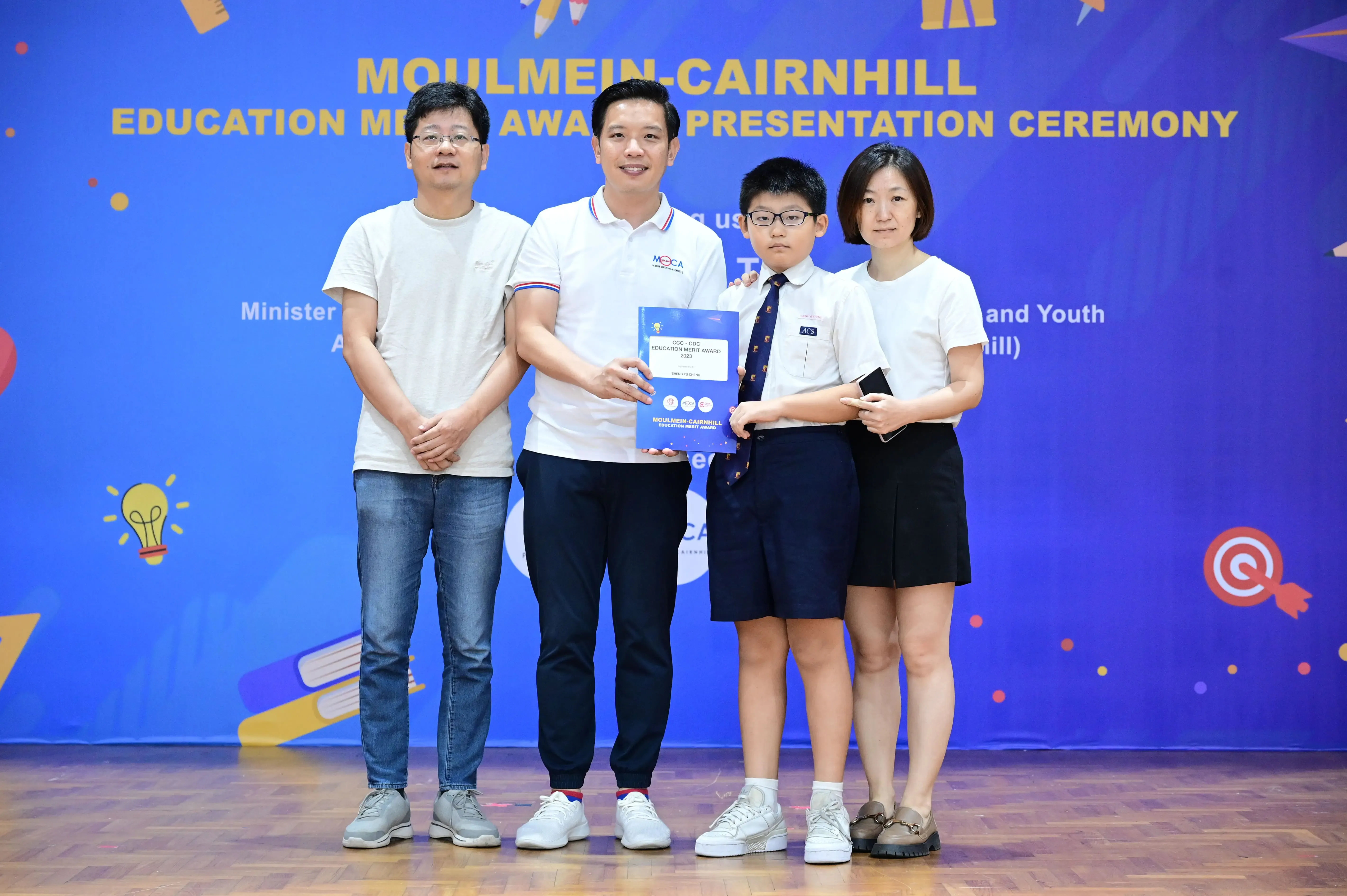 Four people pose at the Moulmein-Cairnhill Education Merit Award Presentation Ceremony, with a student holding an award booklet.