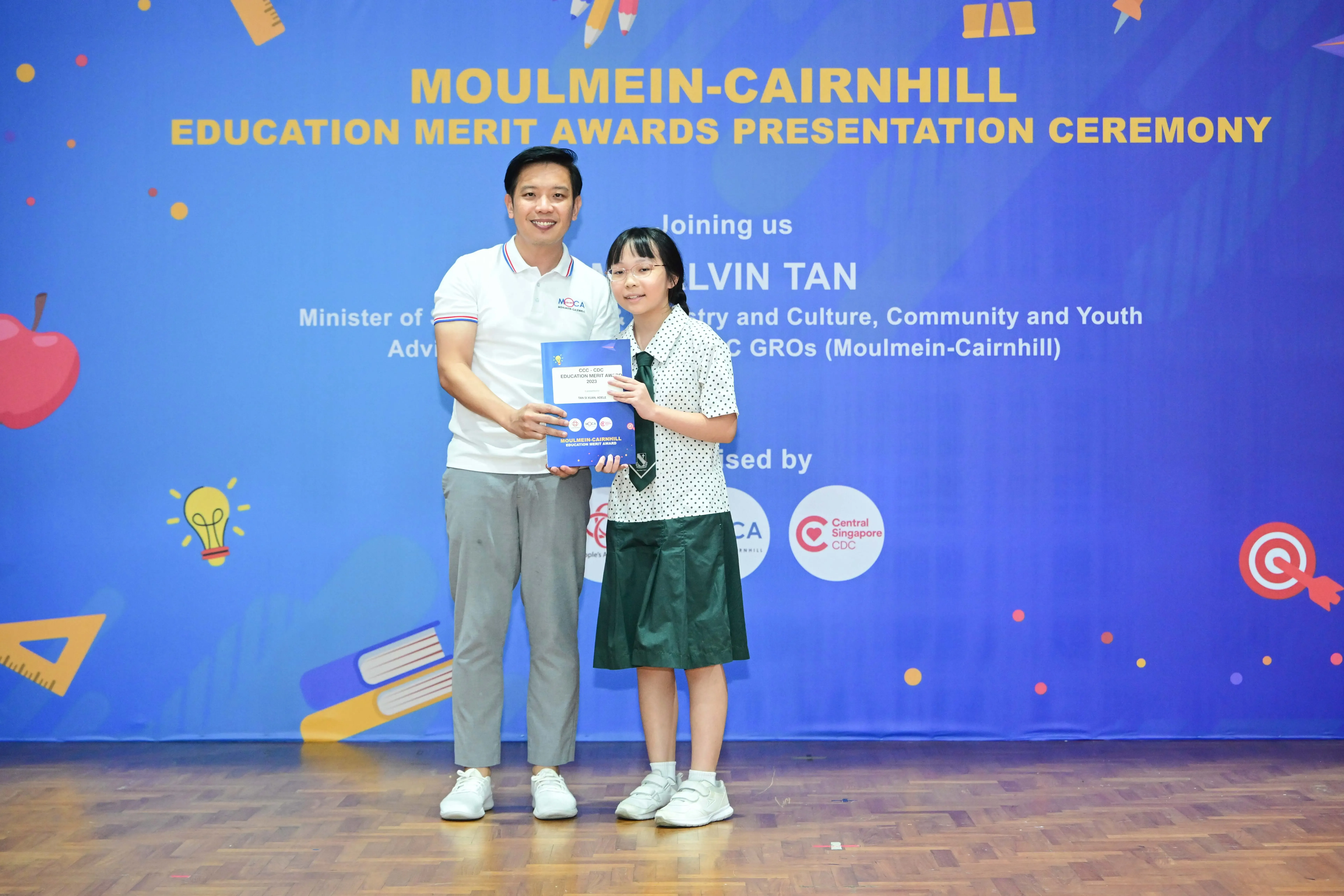 A man and a young girl in school uniform hold an award together at the Moulmein-Cairnhill Education Merit Awards ceremony.