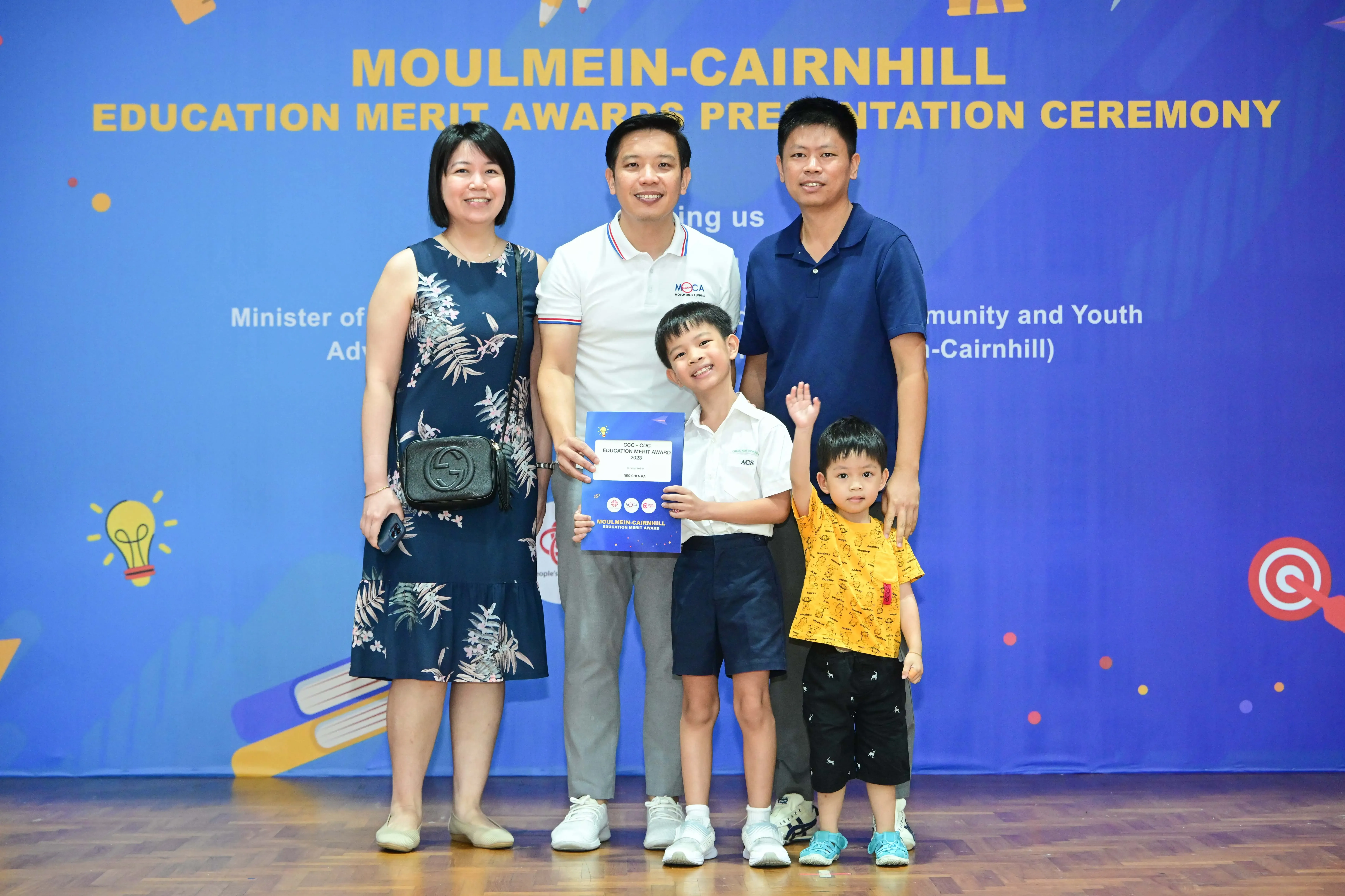 Five people pose in front of a blue Moulmein-Cairnhill Education Merit Awards banner, with a boy holding a certificate.