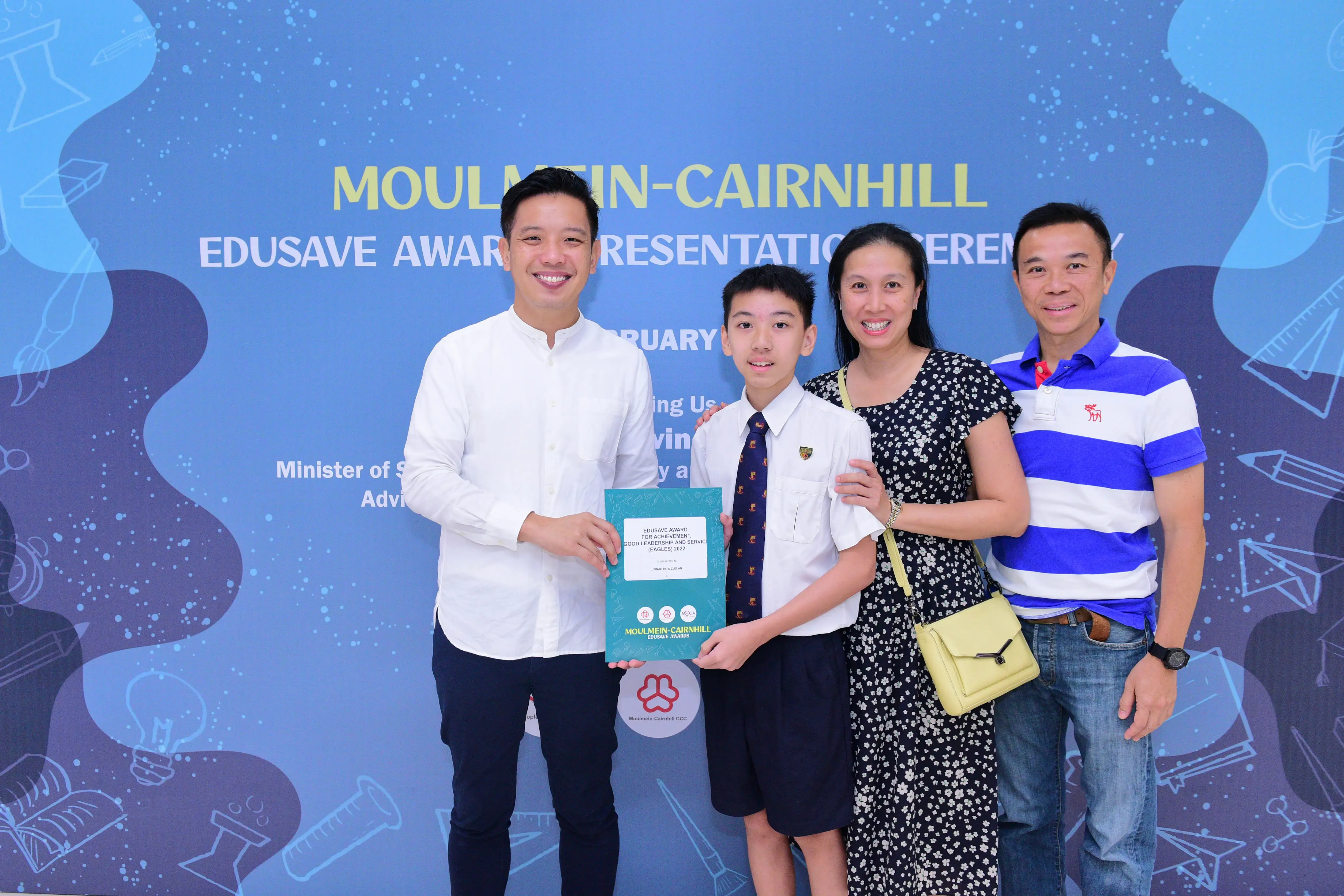 Four people smile at the Moulmein-Cairnhill Edusave Award Presentation Ceremony, with a boy holding a teal award folder.