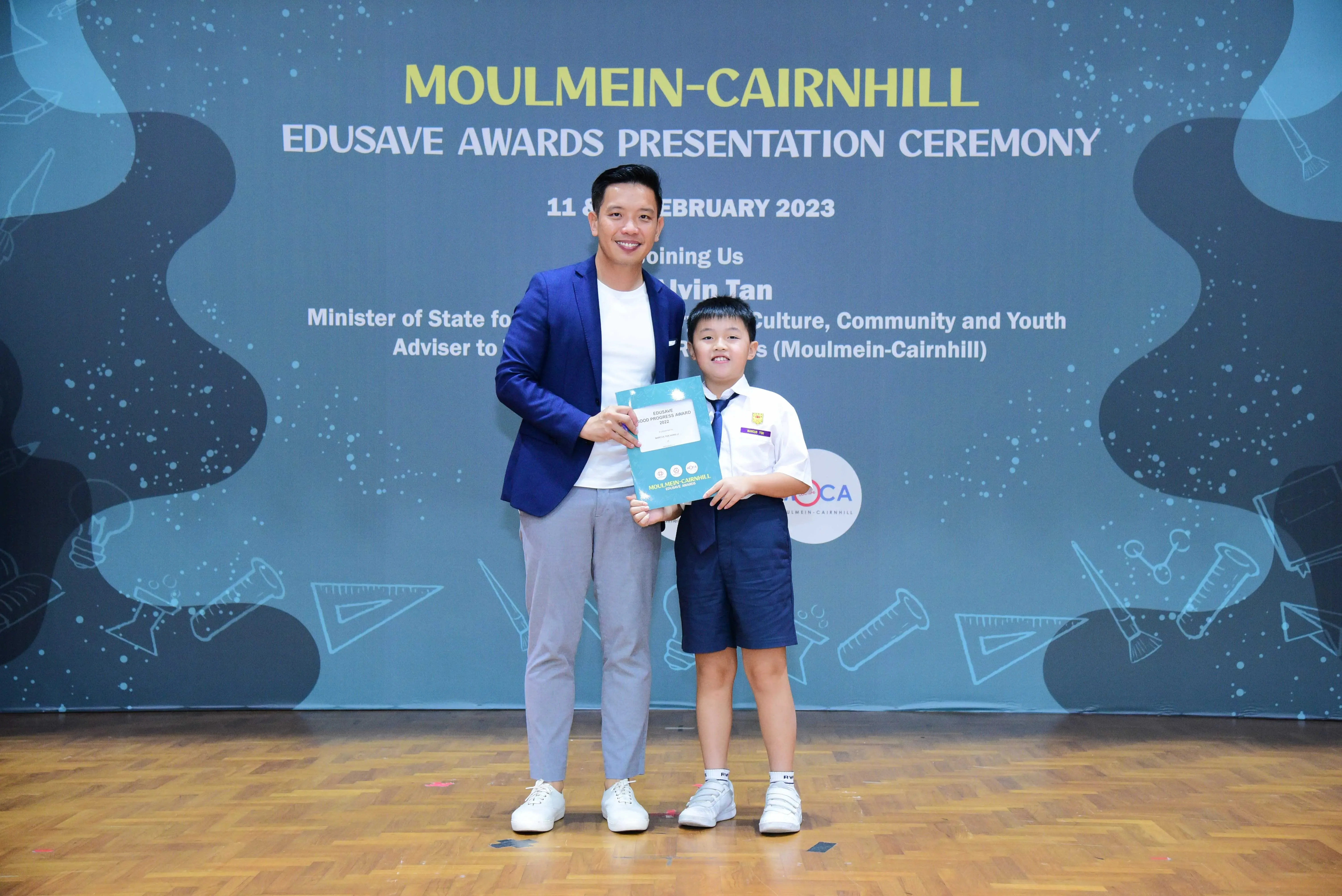 A man in a blue blazer and a young boy in school uniform hold a certificate at the Moulmein-Cairnhill Edusave Awards 2023.
