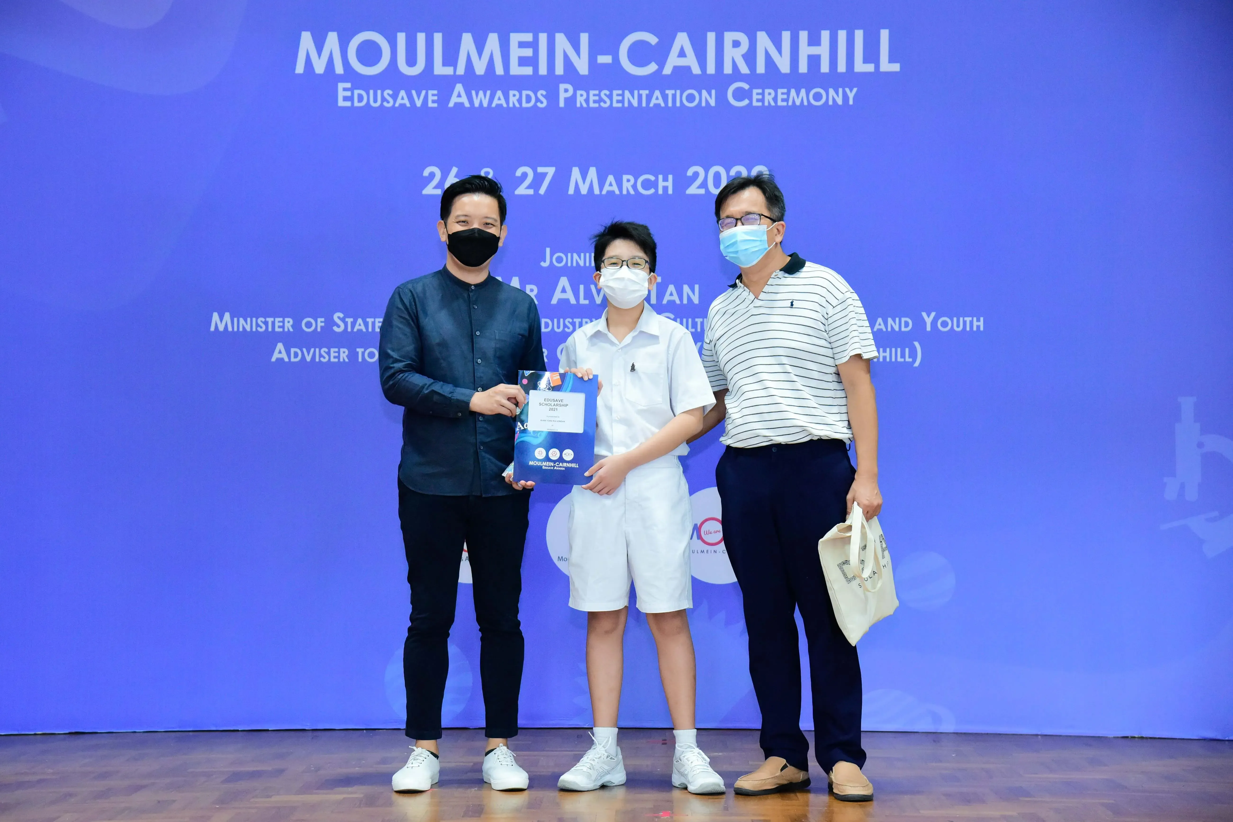 Three masked people pose at the Moulmein-Cairnhill Edusave Awards Presentation Ceremony, with a student holding an award.