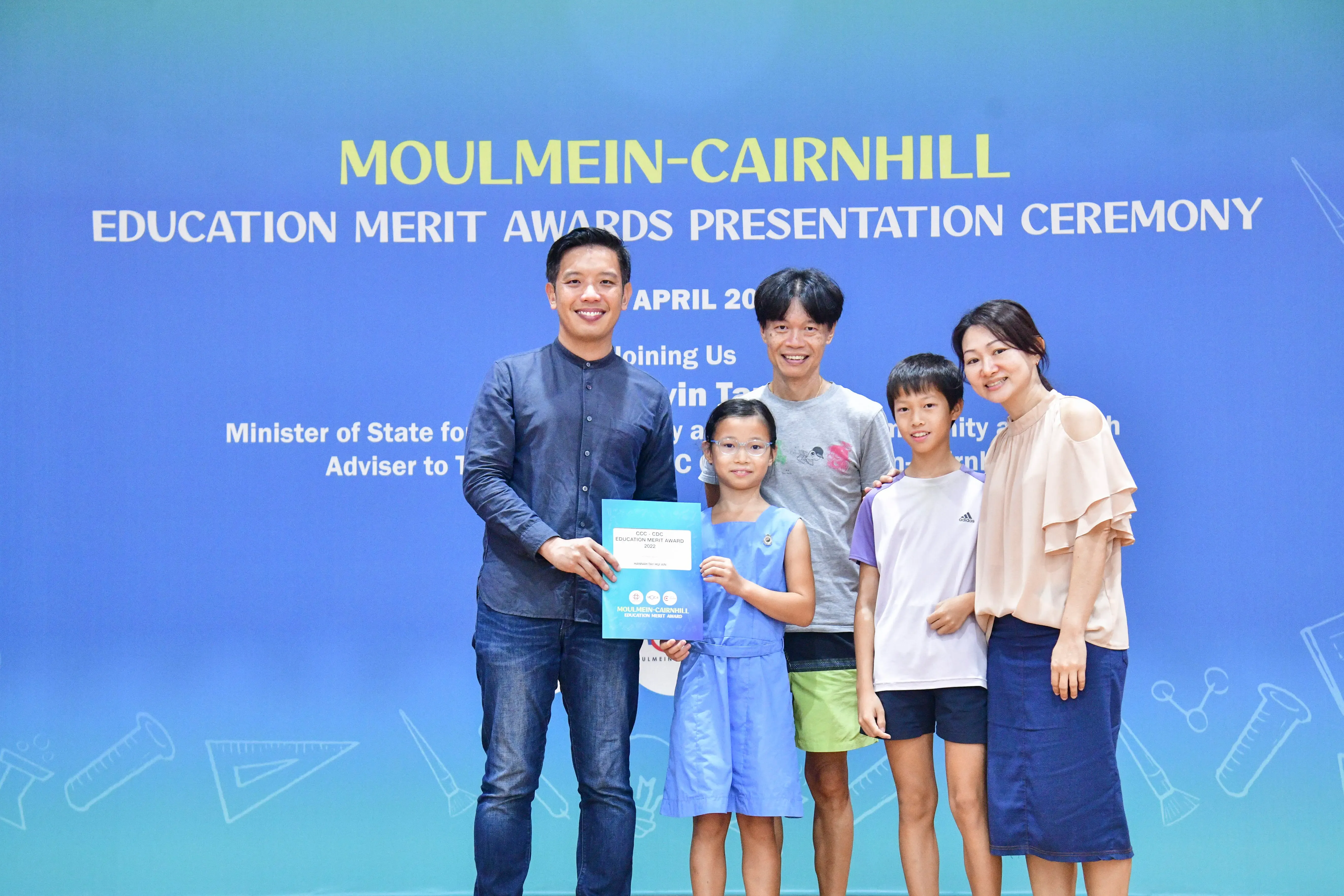 Five people pose before a Moulmein-Cairnhill Education Merit Awards banner; a girl holds a blue award certificate.