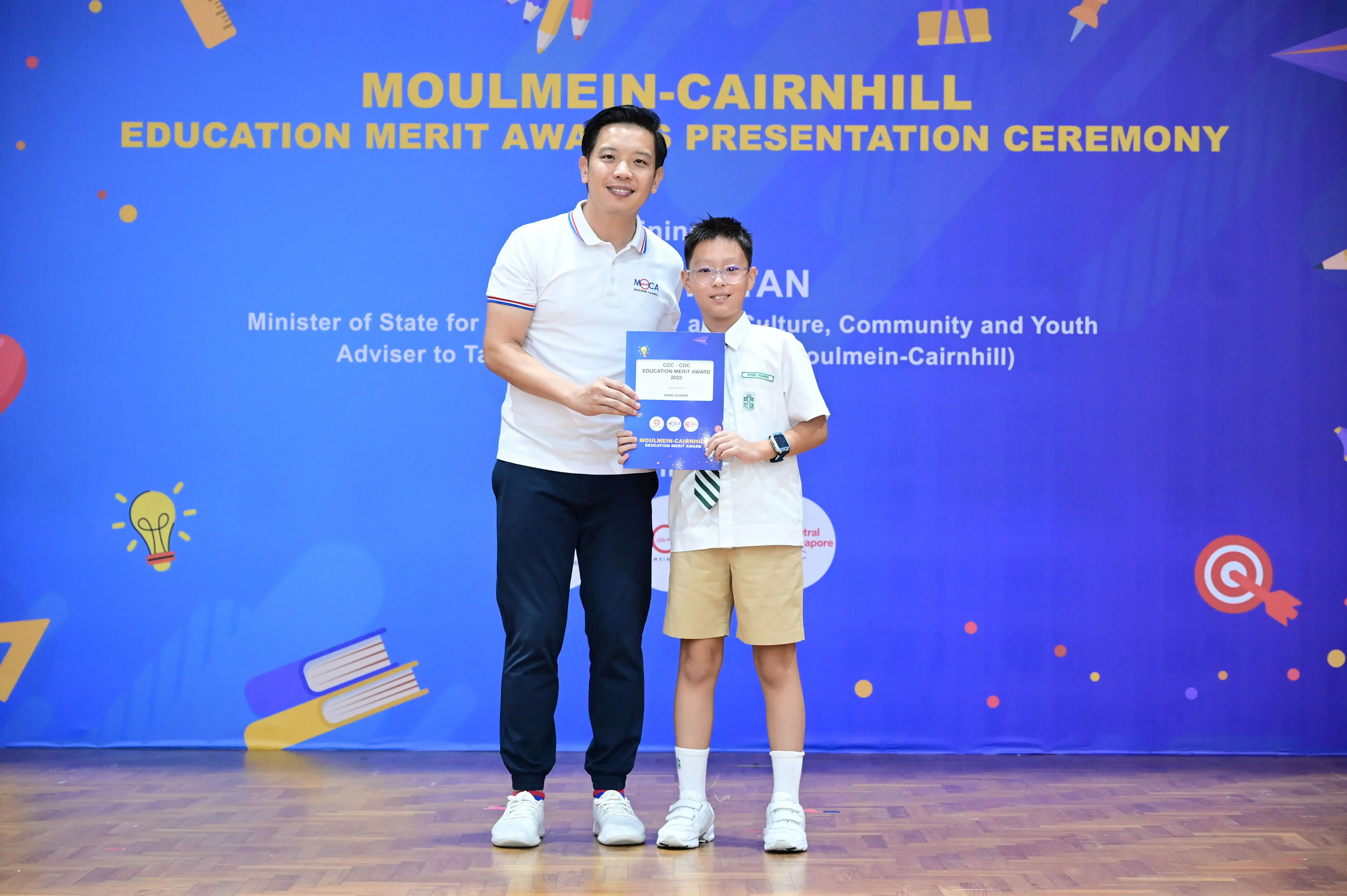 An adult and a young boy holding an award certificate at the Moulmein-Cairnhill Education Merit Awards ceremony.
