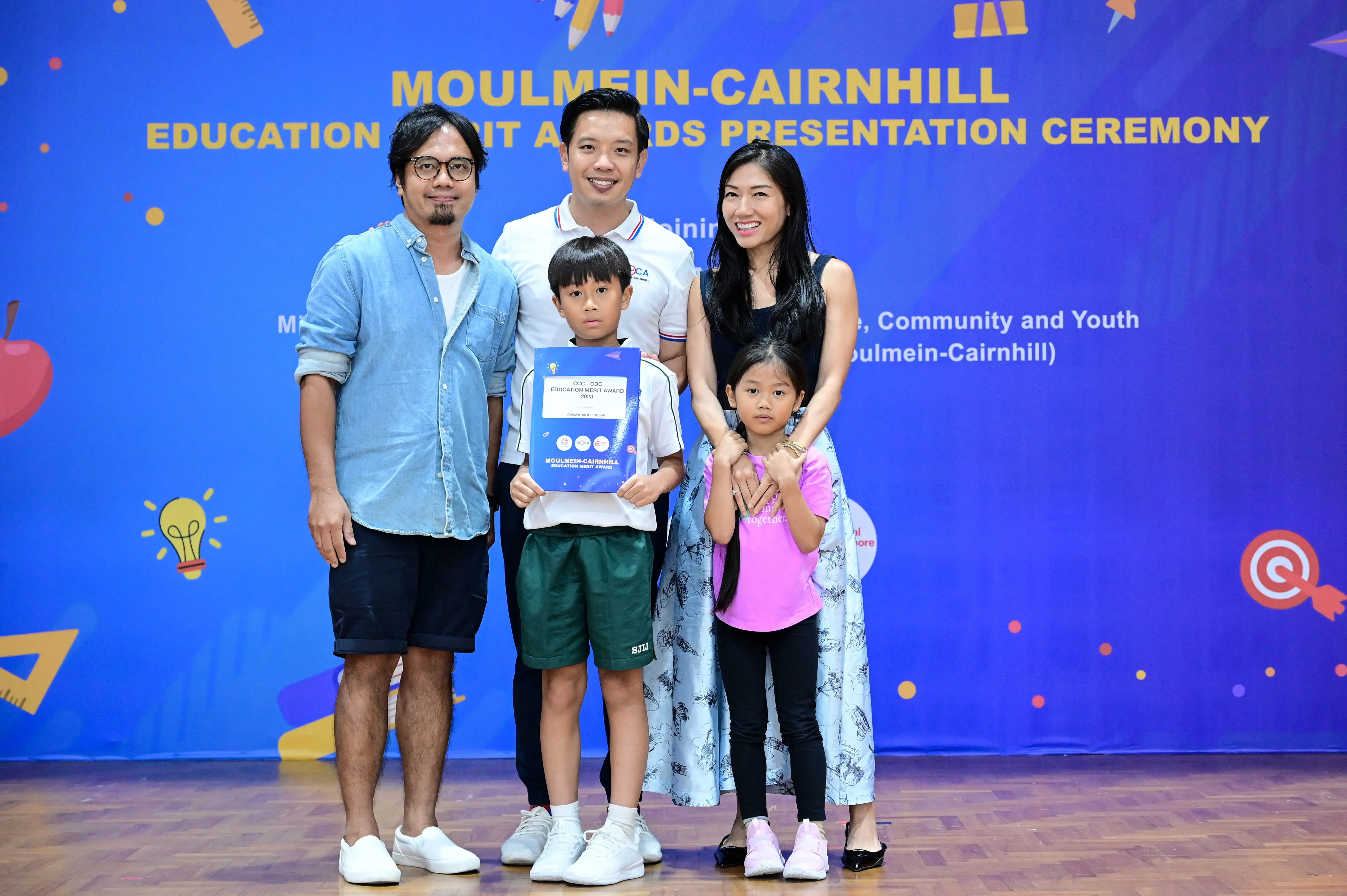Five people pose in front of a blue Moulmein-Cairnhill Education Merit Awards banner, with a boy holding a certificate.