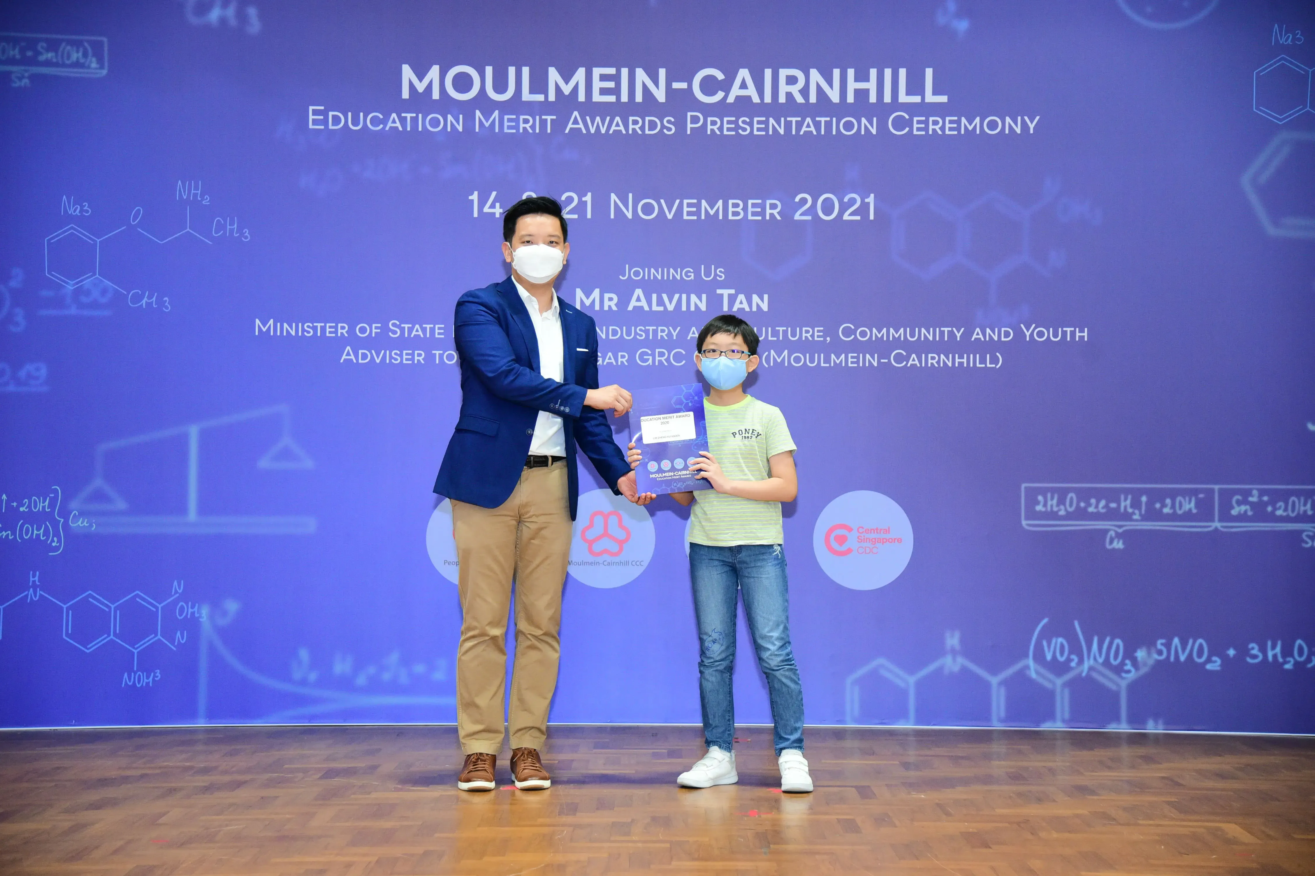 An adult in a blue blazer presents an award to a young boy on stage at the Moulmein-Cairnhill Education Merit Awards 2021.