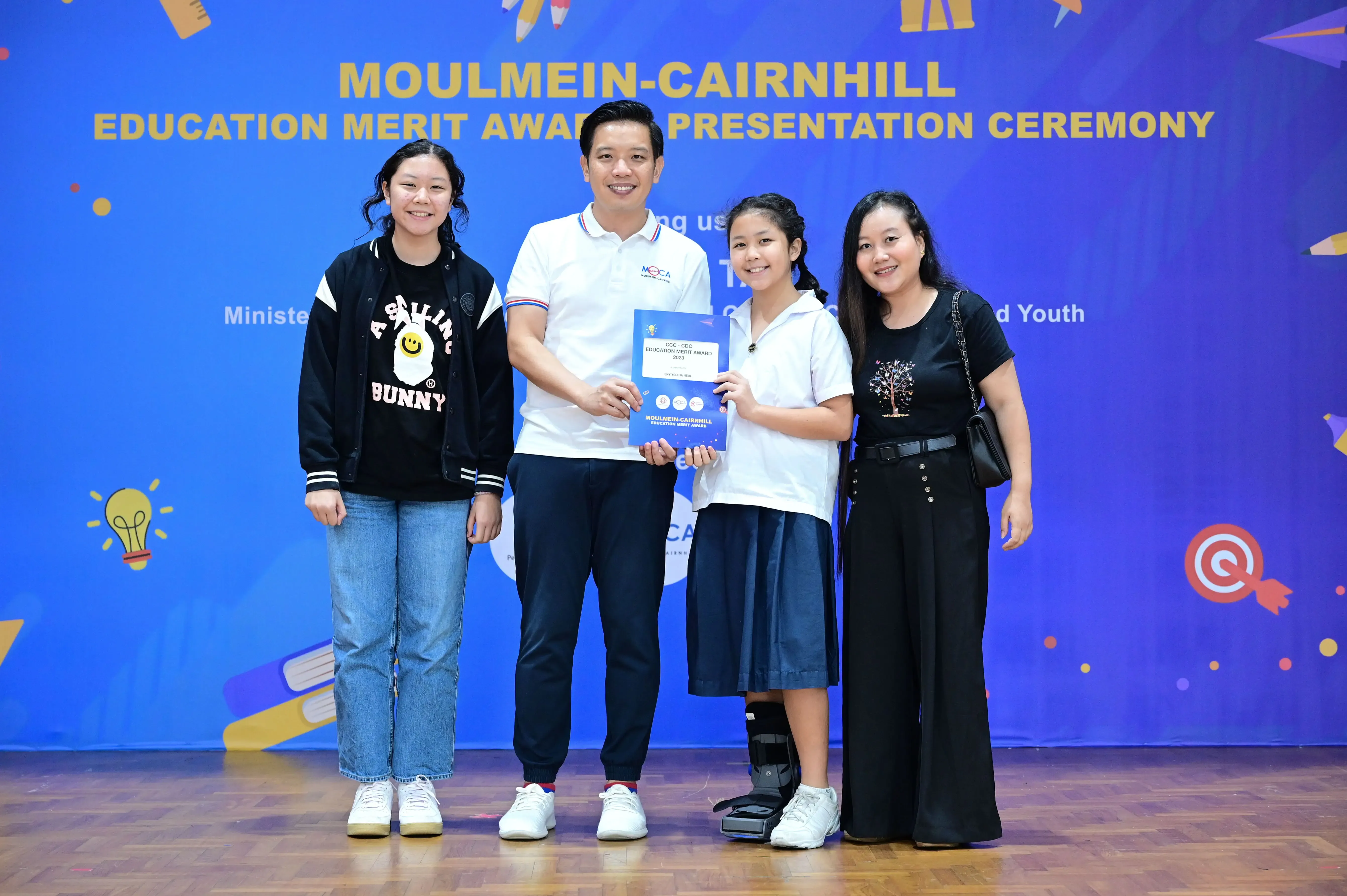 Four people pose at the Moulmein-Cairnhill Education Merit Award Presentation Ceremony, with a student holding an award certificate.