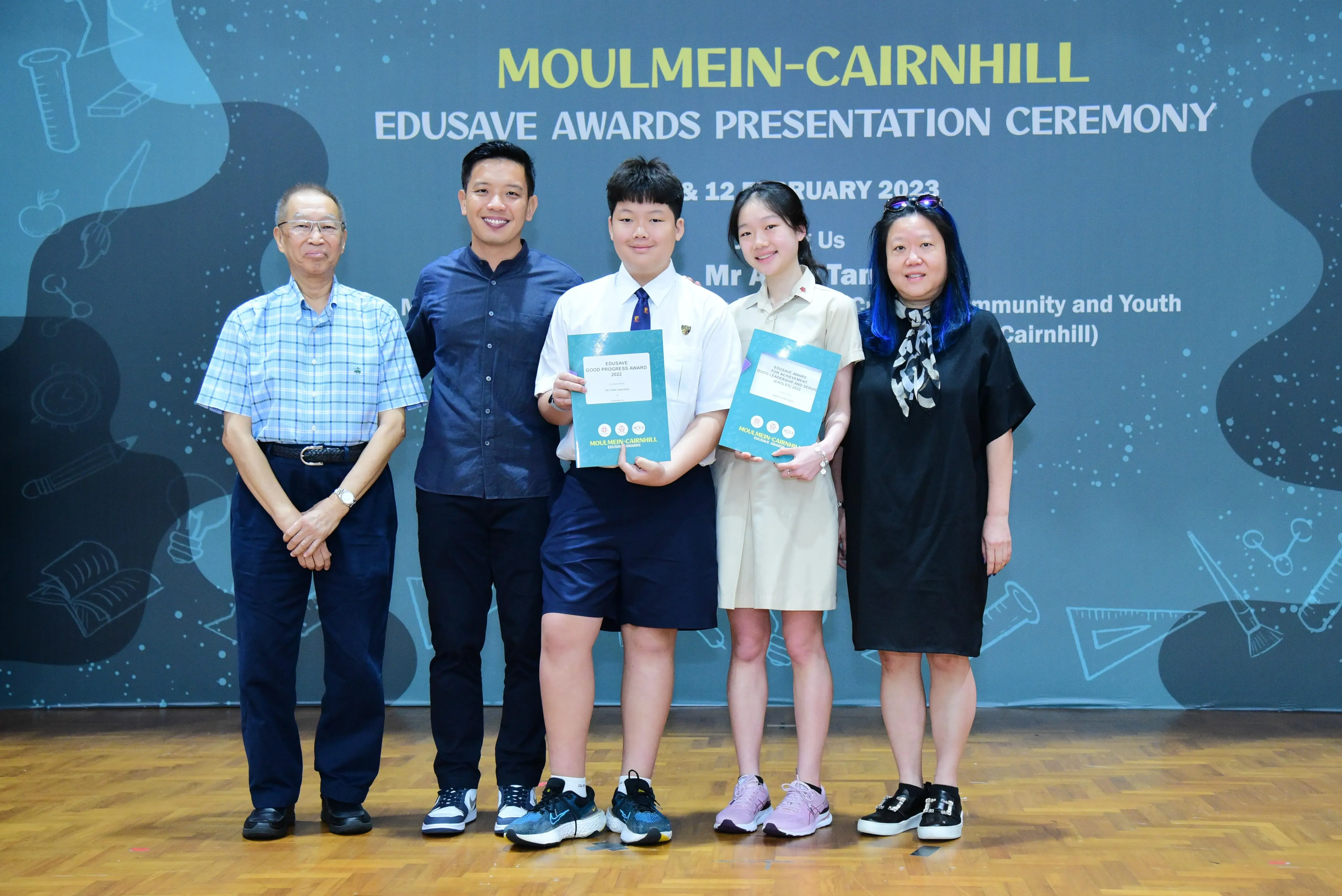 Five people pose at the Moulmein-Cairnhill Edusave Awards Presentation Ceremony, two students holding teal award booklets.