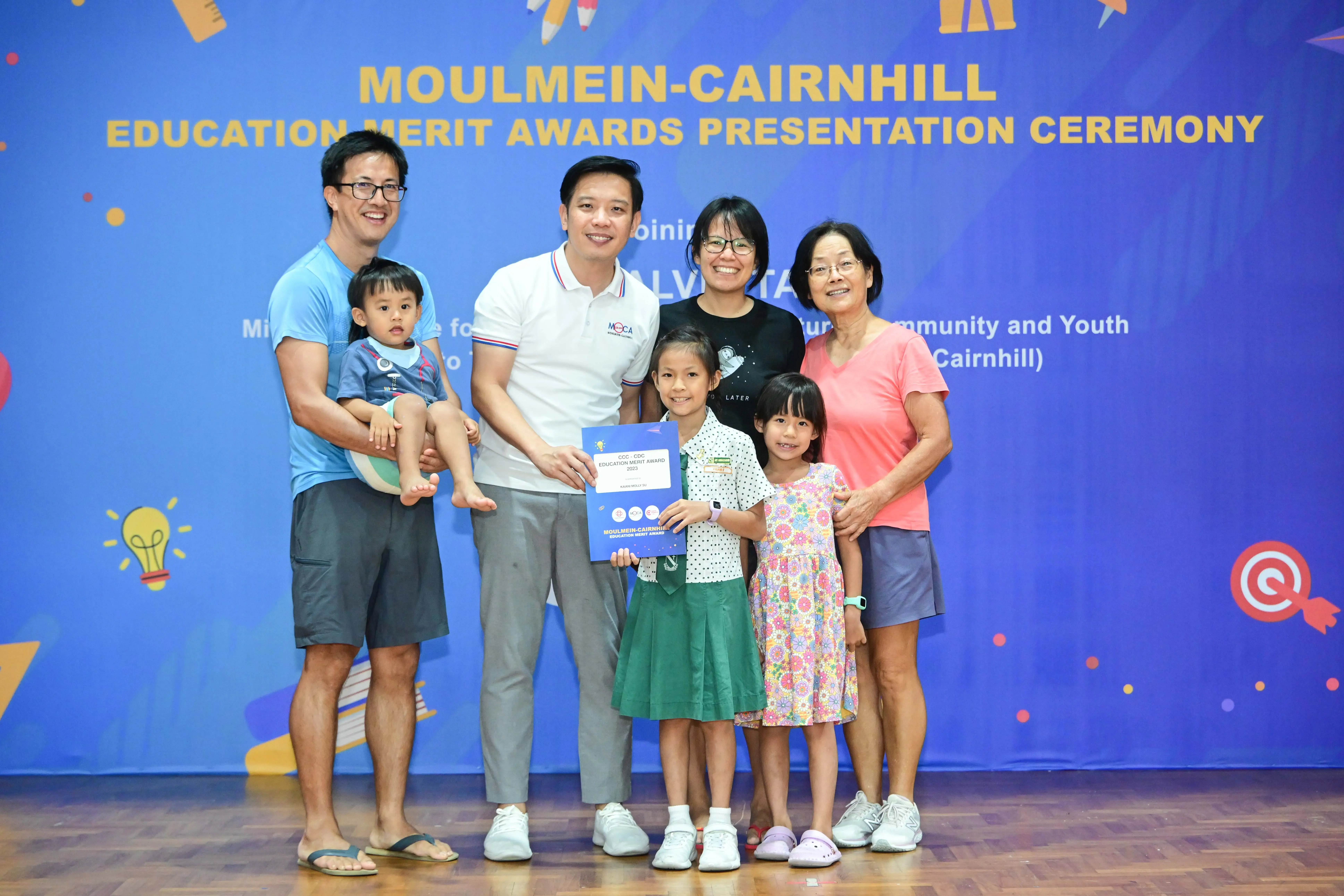 Family of seven posing with an award certificate at the Moulmein-Cairnhill Education Merit Awards Presentation Ceremony.