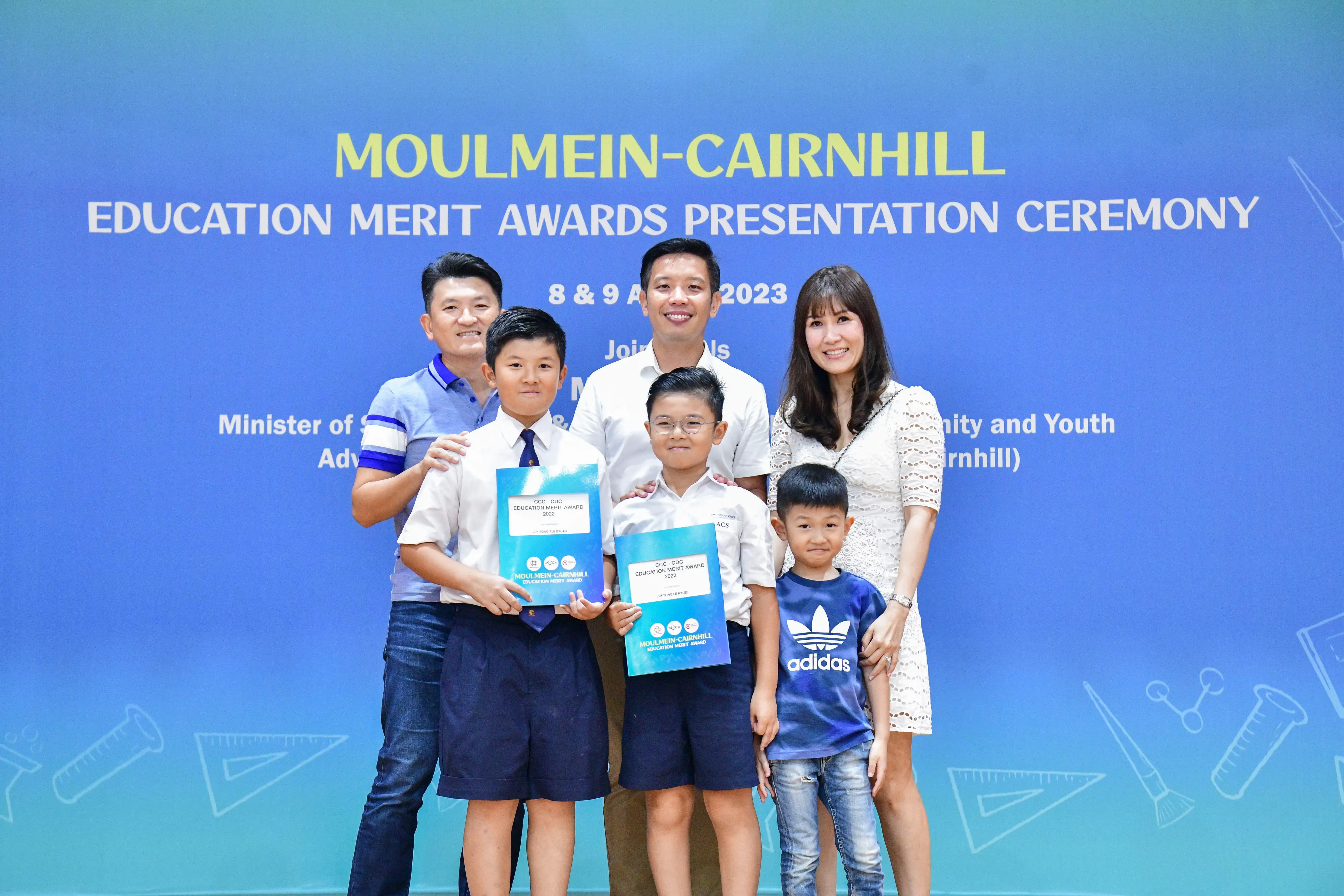 Family of six posing at the Moulmein-Cairnhill Education Merit Awards Ceremony 2023, two boys holding blue award certificates.