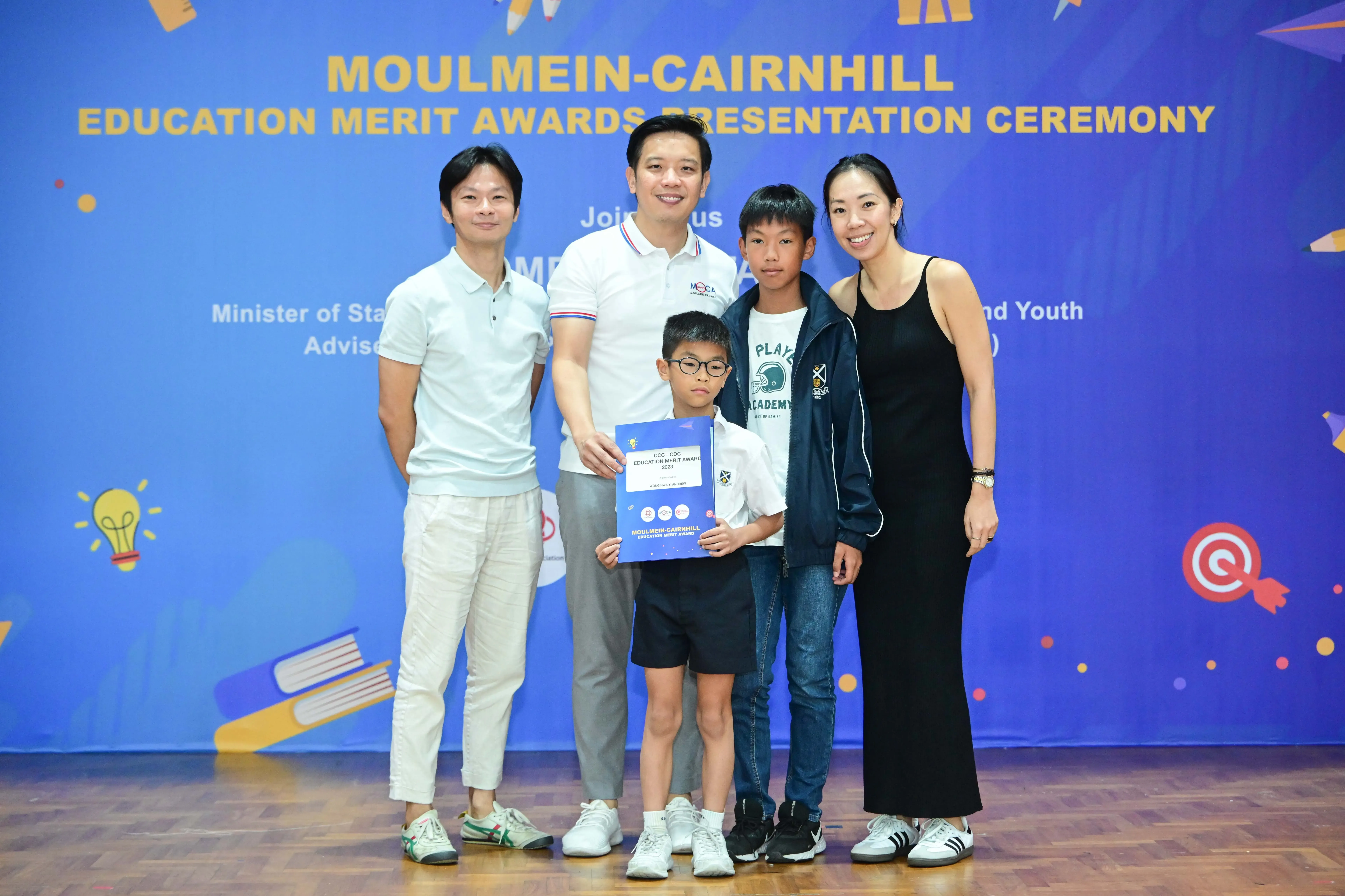 Five people pose at the Moulmein-Cairnhill Education Merit Awards Presentation Ceremony, with a boy holding a certificate.