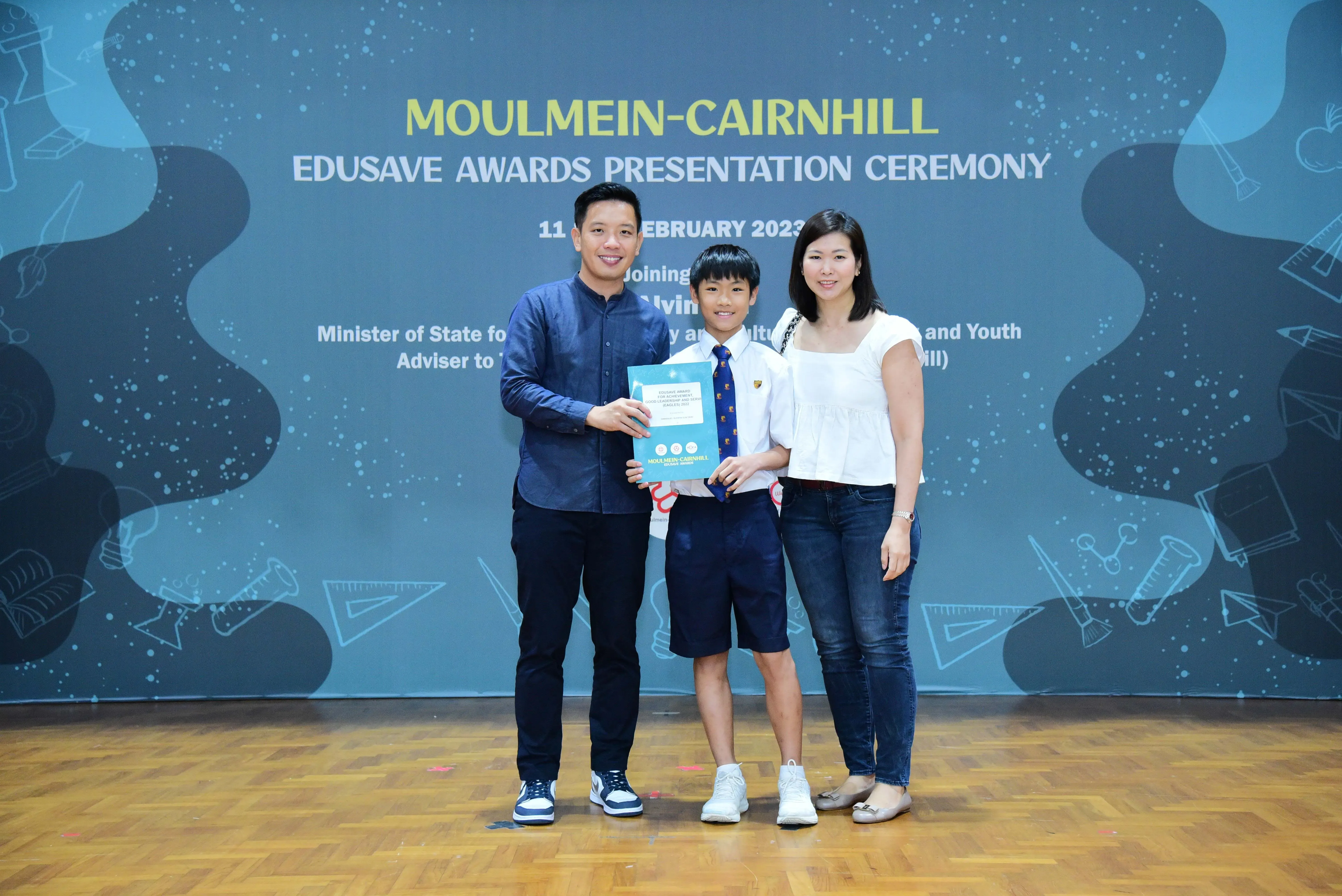 A boy in school uniform holds an award booklet flanked by two adults at the Moulmein-Cairnhill Edusave Awards Ceremony 2023.