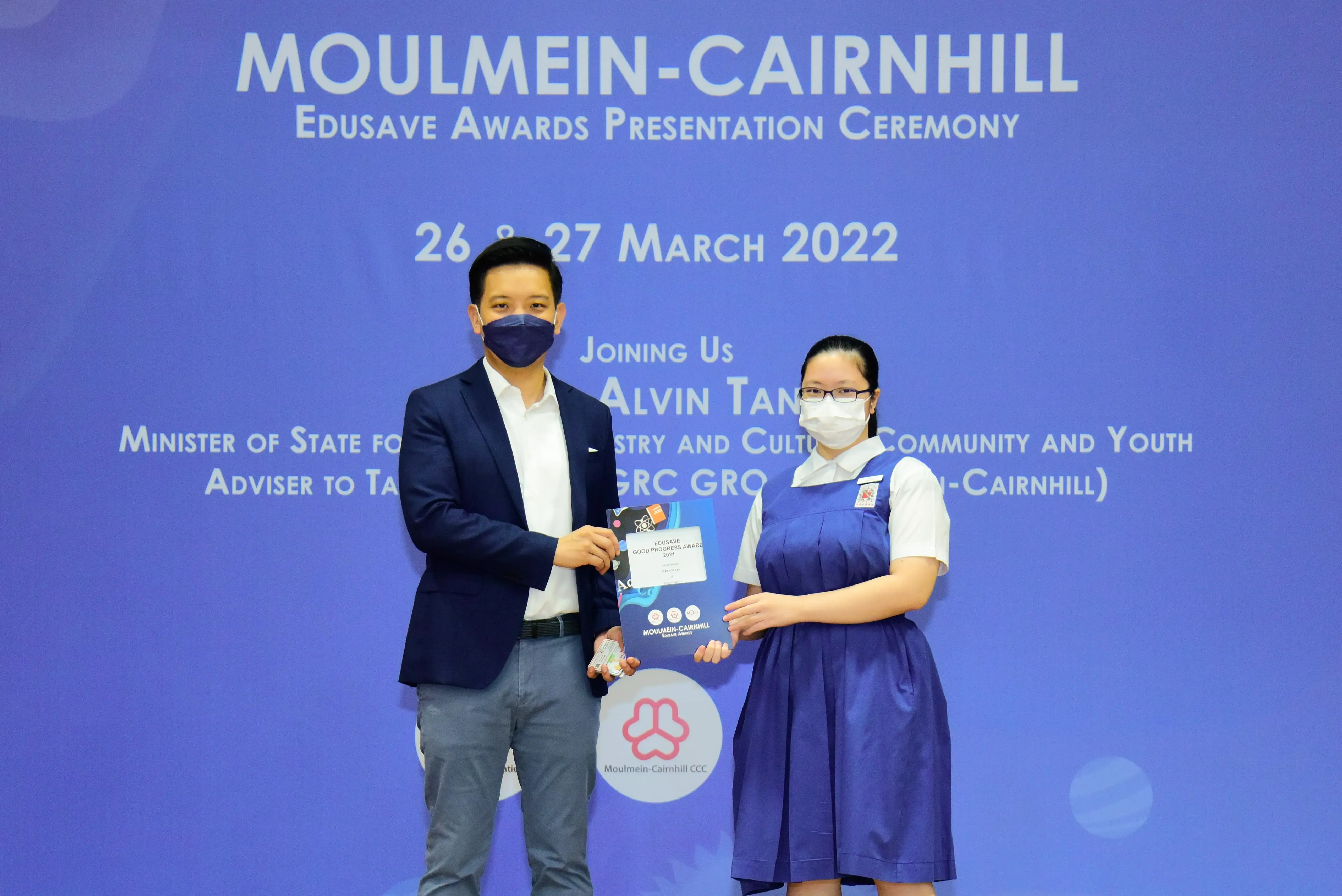A man in a blazer and a girl in school uniform hold an award certificate at the Moulmein-Cairnhill Edusave ceremony.