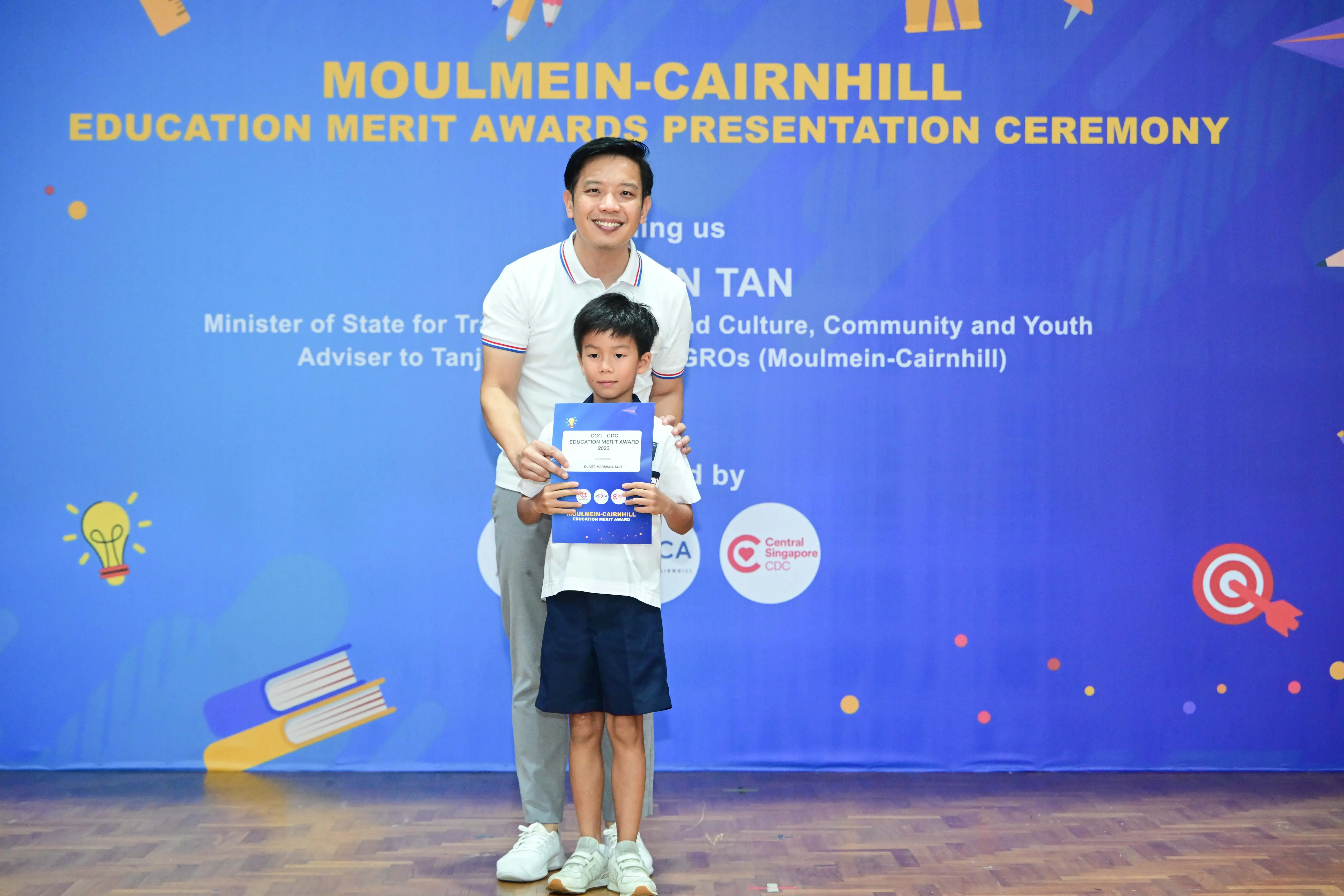 A man and young boy hold a blue certificate at the Moulmein-Cairnhill Education Merit Awards Presentation Ceremony.
