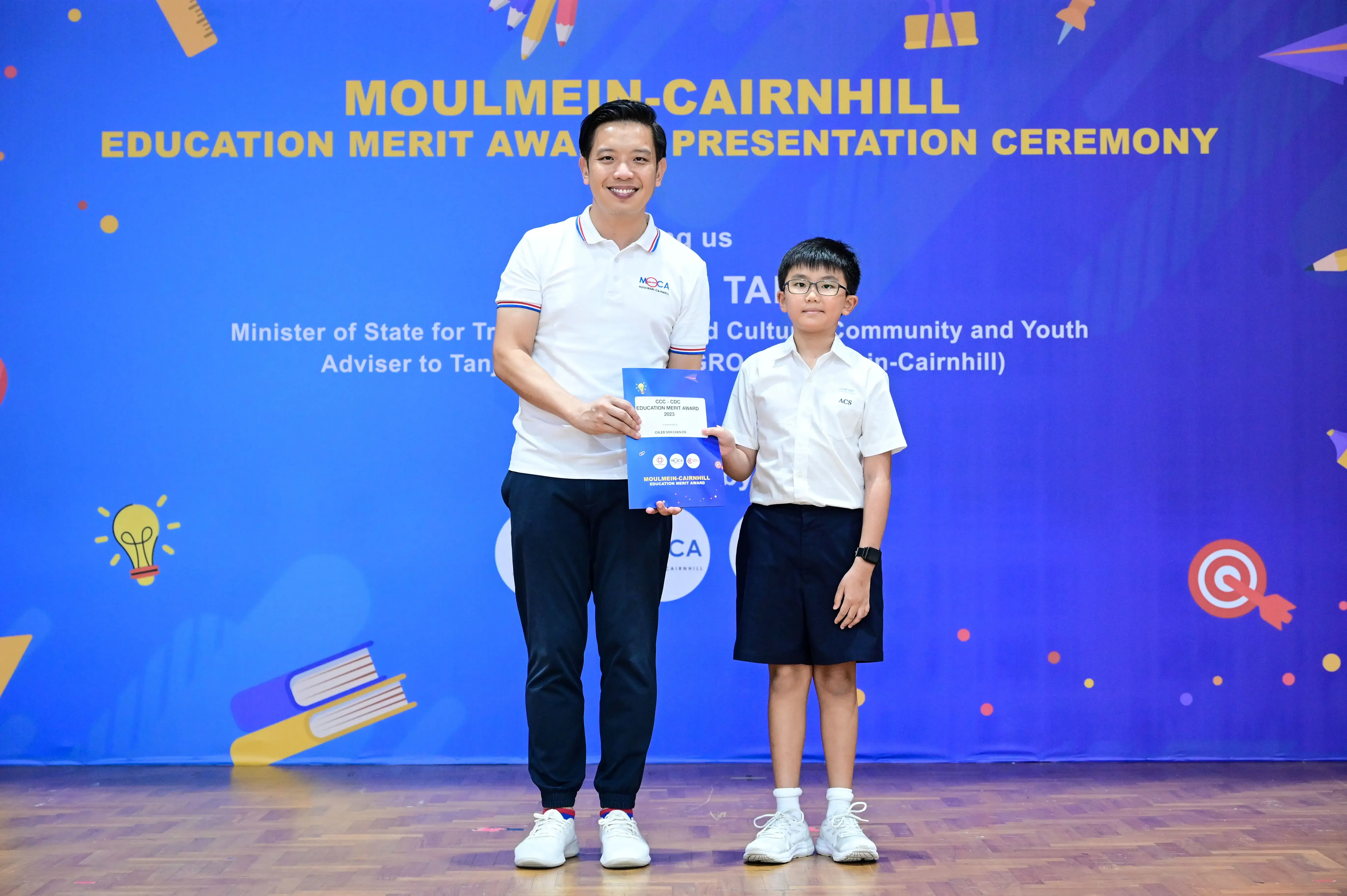 A man and a young boy hold a blue certificate at the Moulmein-Cairnhill Education Merit Award Presentation Ceremony.