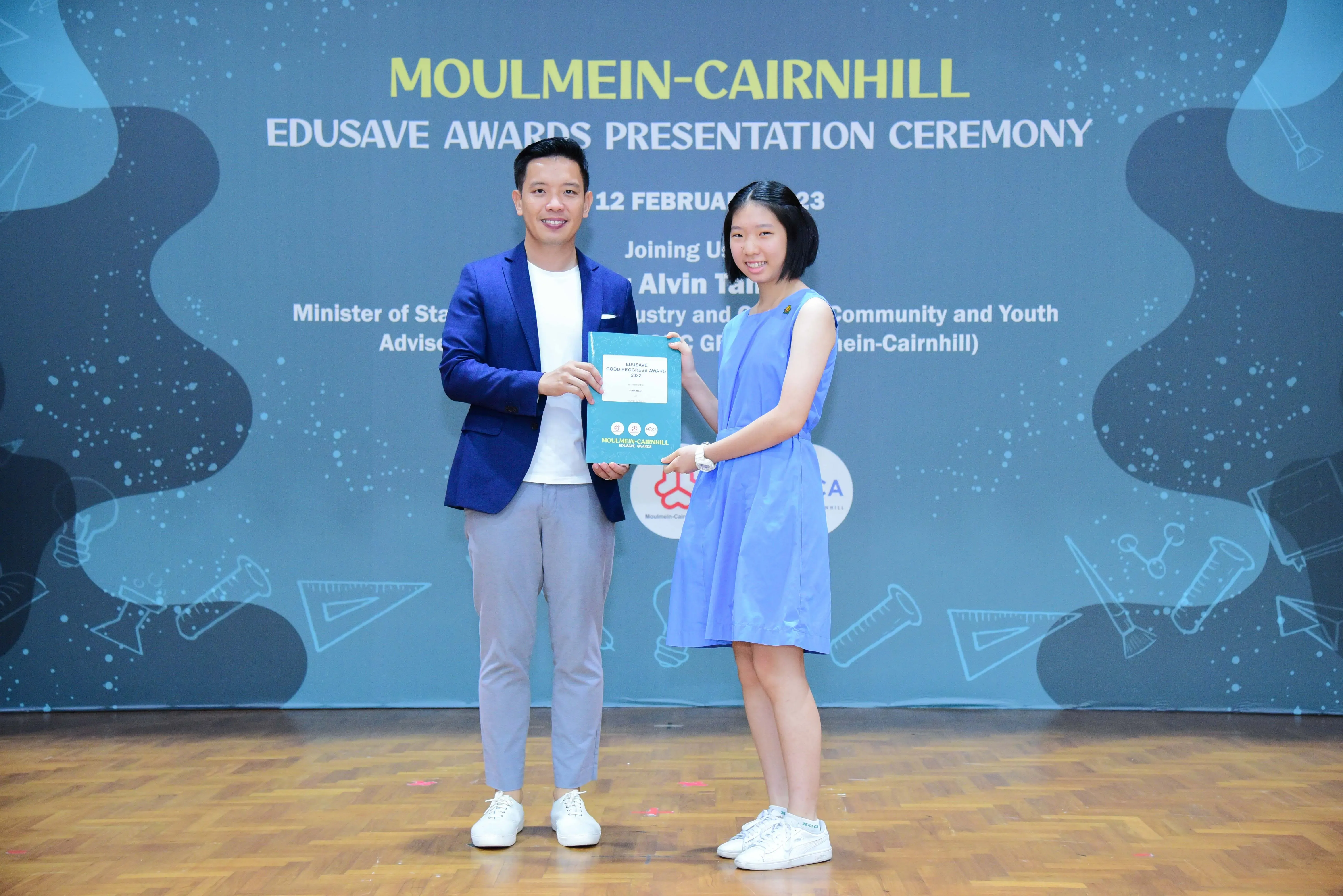 A man in a blue blazer and a girl in a blue dress hold an award together at the Moulmein-Cairnhill Edusave Awards Ceremony.