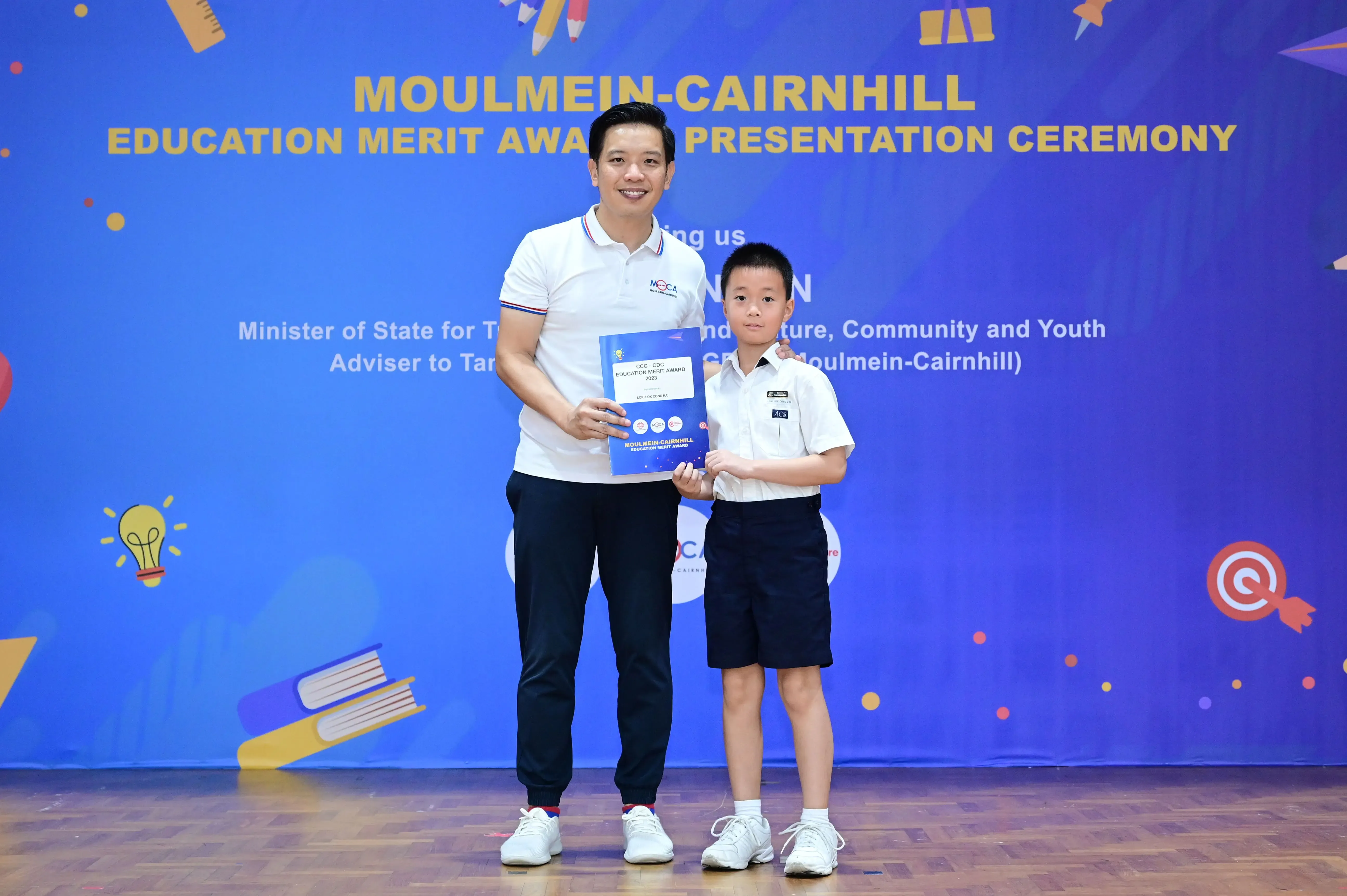 Adult and young boy hold a blue award booklet at Moulmein-Cairnhill Education Merit Award Presentation Ceremony.
