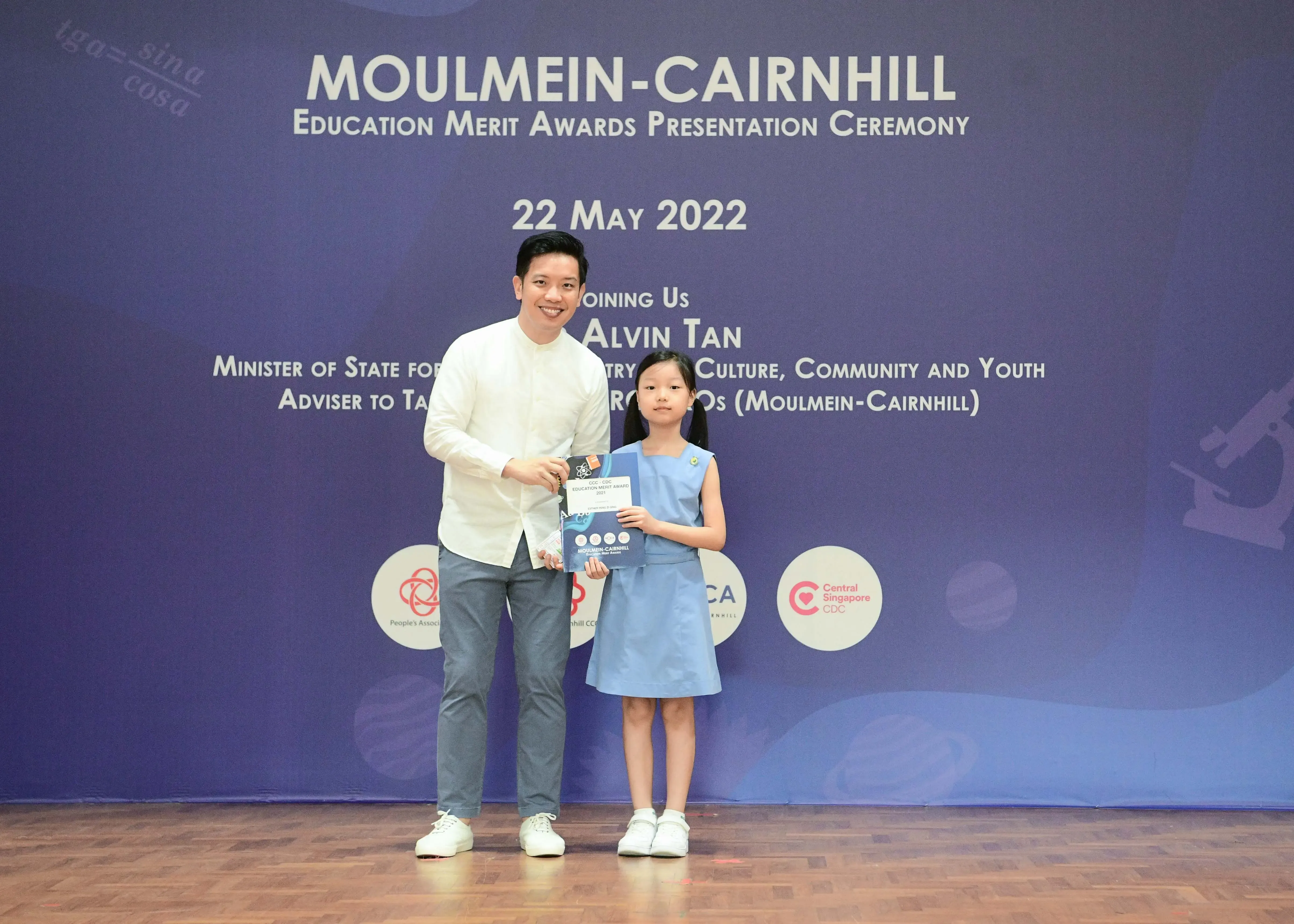 A man and a young girl holding an award certificate on stage at the Moulmein-Cairnhill Education Merit Awards ceremony.