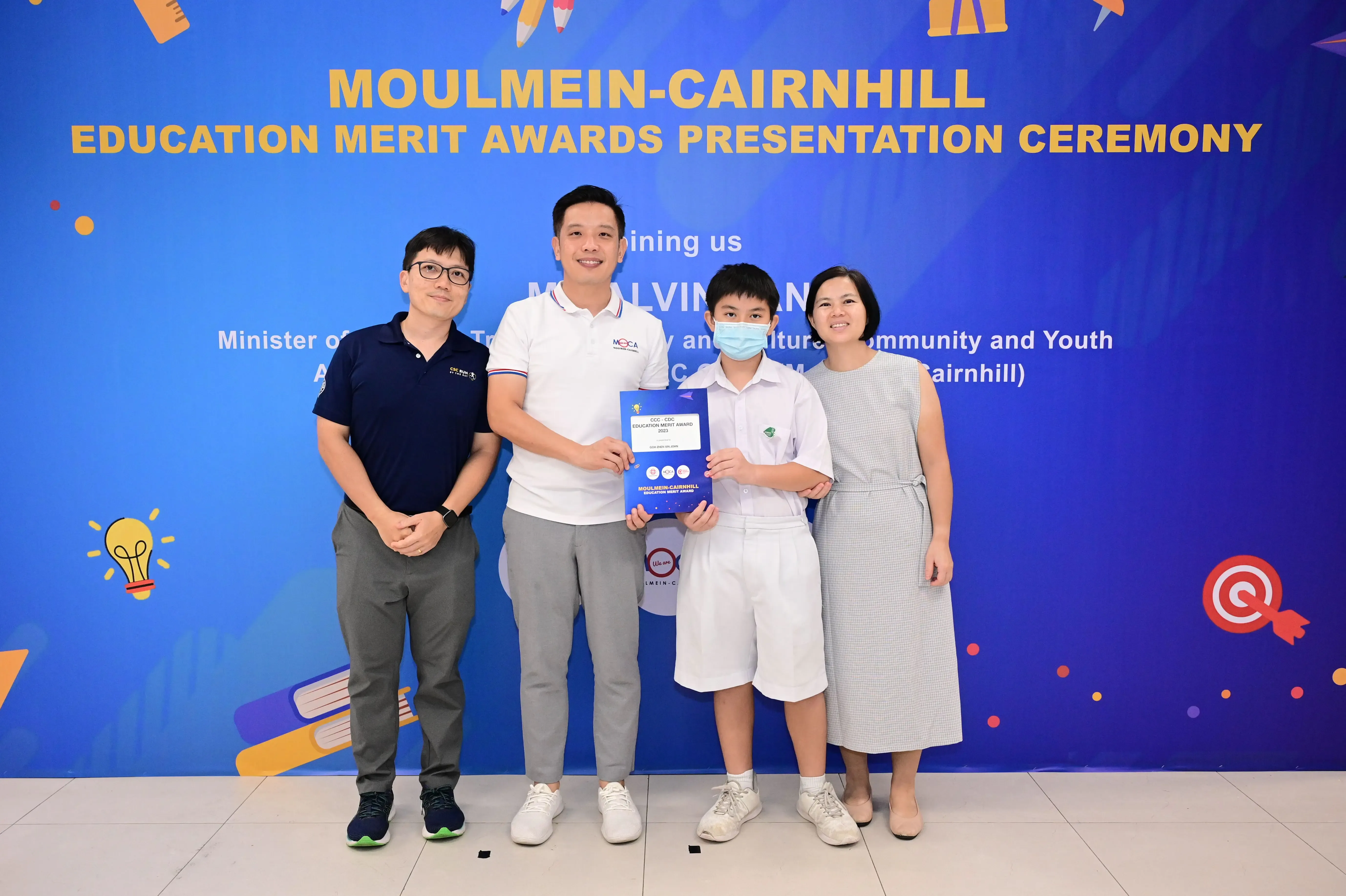 Four people pose before a Moulmein-Cairnhill Education Merit Awards banner, with a boy holding a blue award certificate.