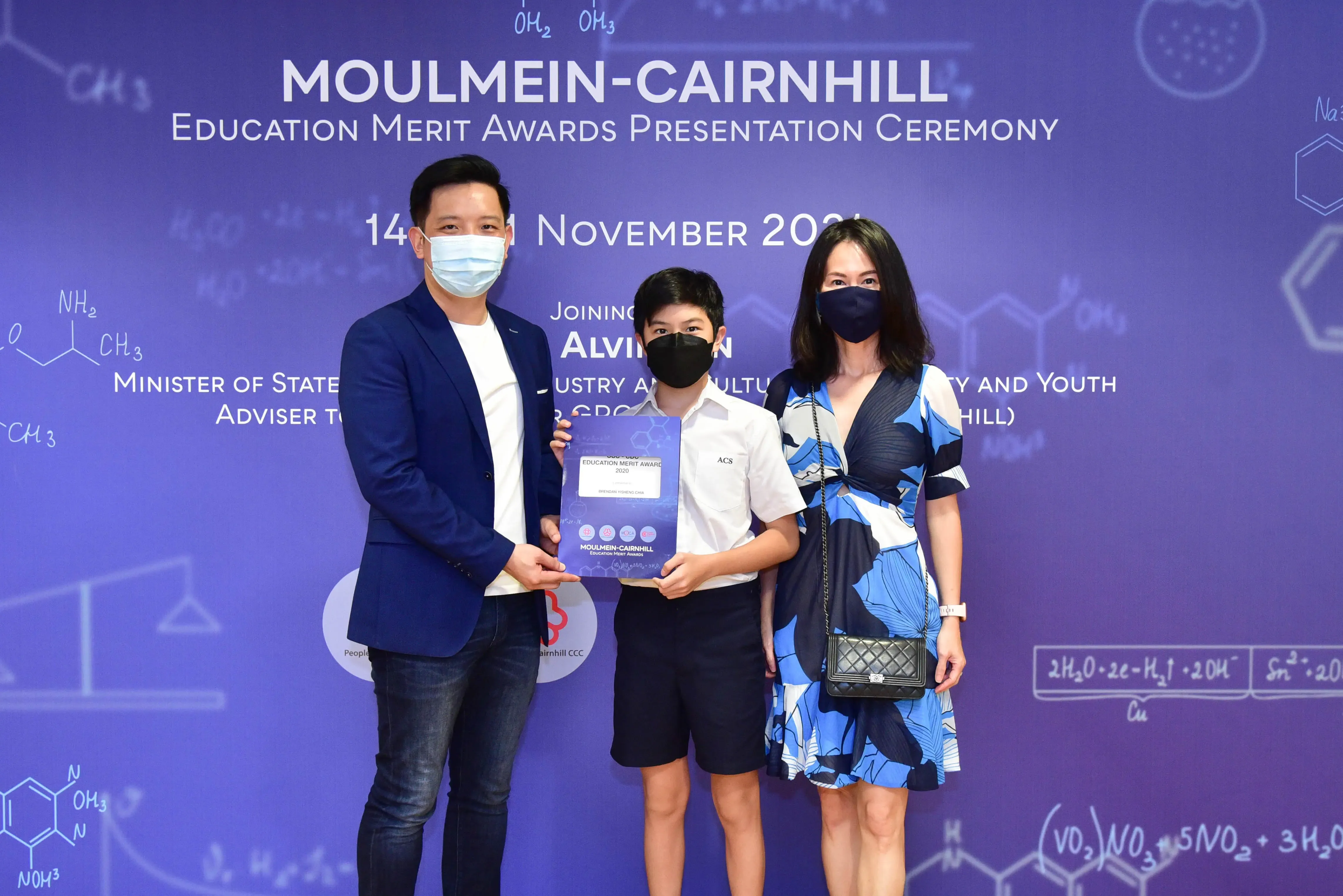 Three masked people pose at the Moulmein-Cairnhill Education Merit Awards Presentation Ceremony, with a boy holding an award.