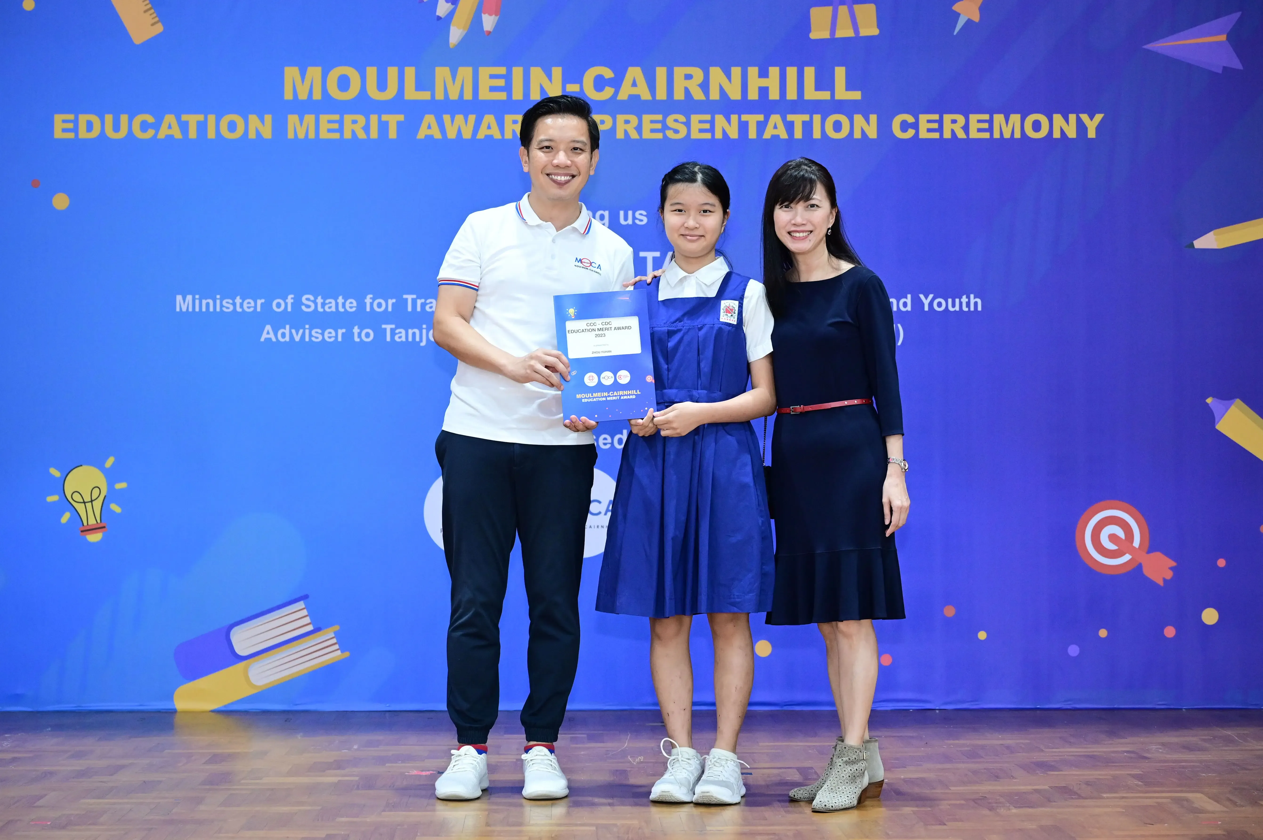 Three people pose together holding a certificate at the Moulmein-Cairnhill Education Merit Award Presentation Ceremony.