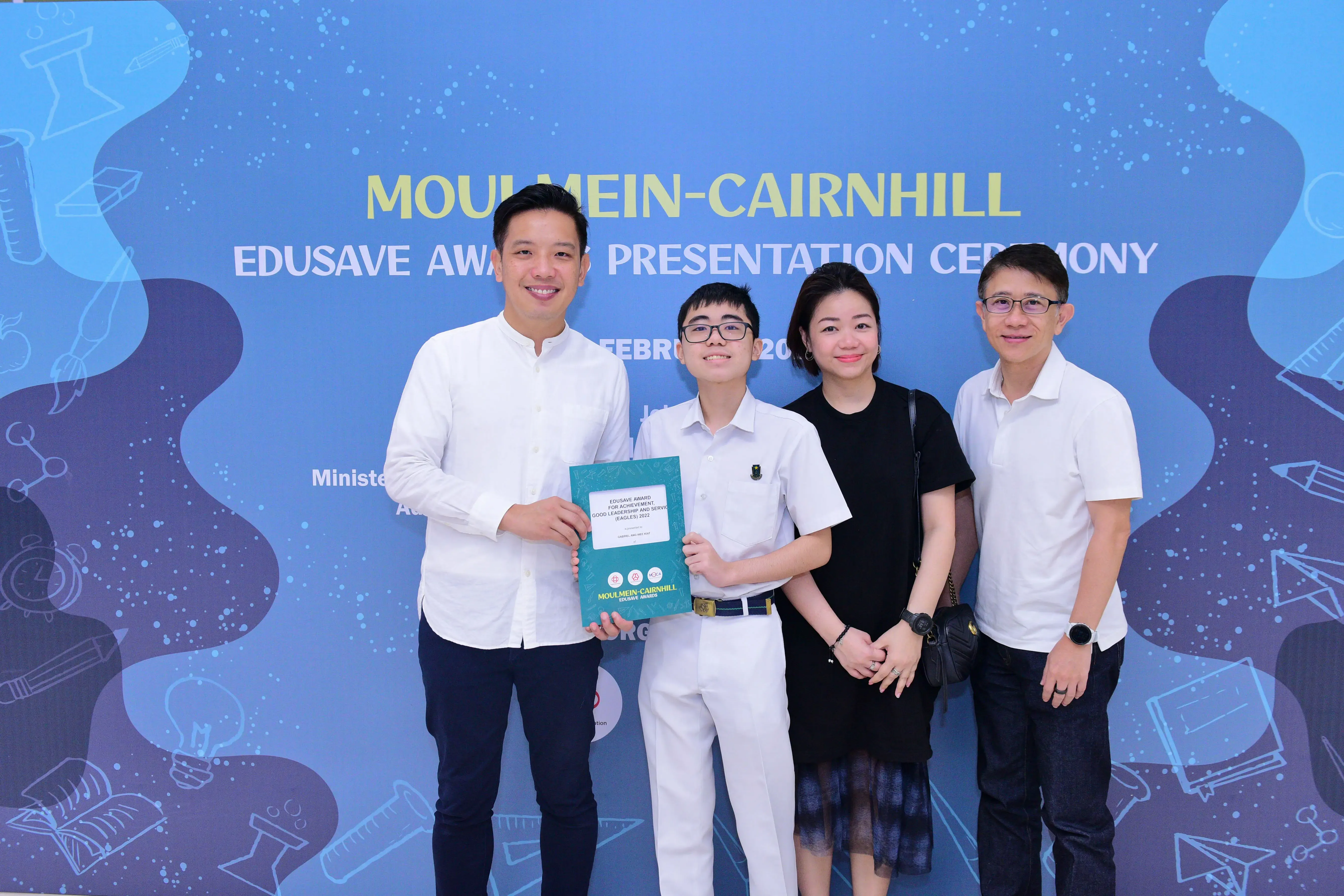 Four people pose smiling at the Moulmein-Cairnhill Edusave Awards Presentation Ceremony, a student holding an award booklet.