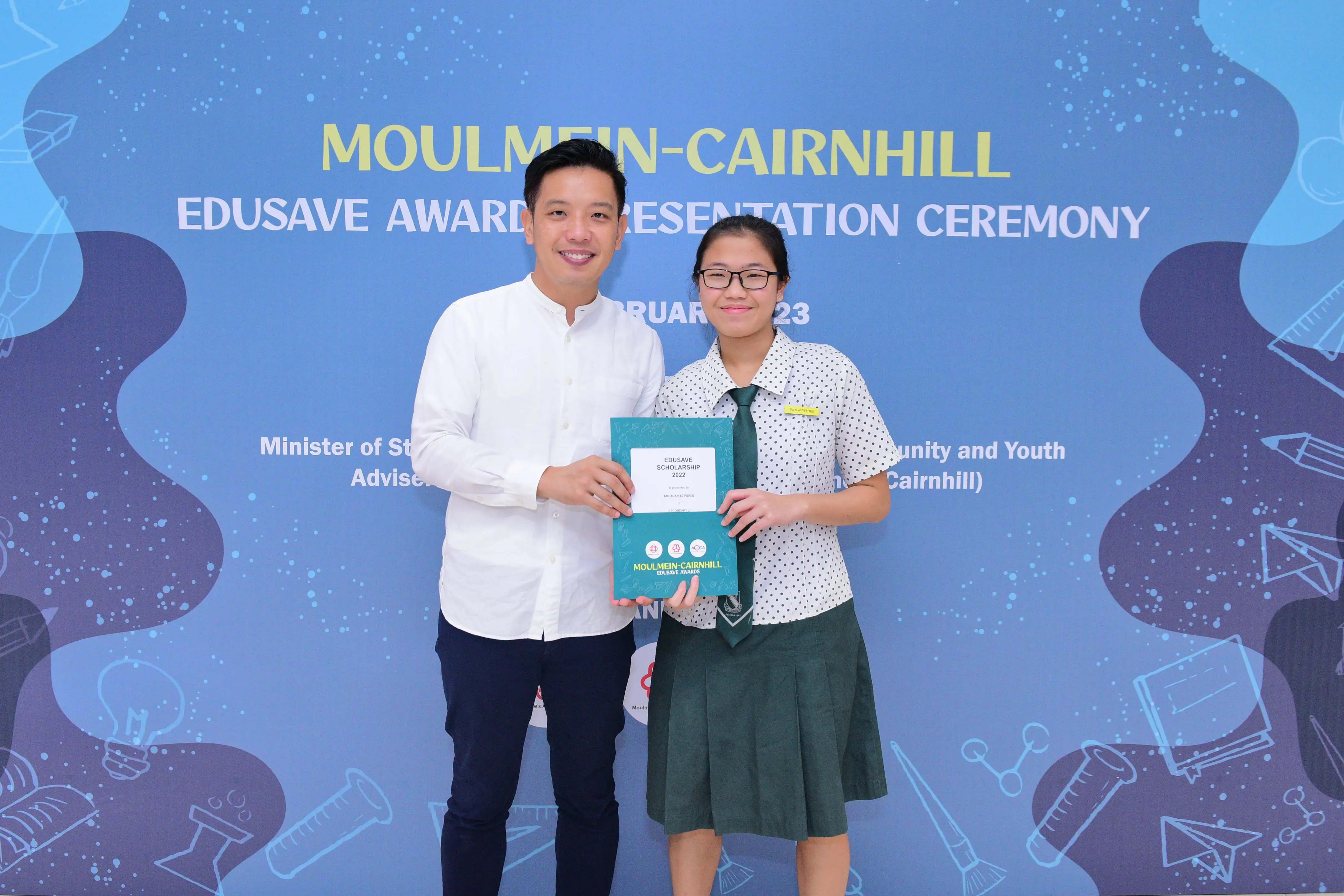 A man and a student in uniform hold an Edusave Scholarship award at the Moulmein-Cairnhill Awards Ceremony.