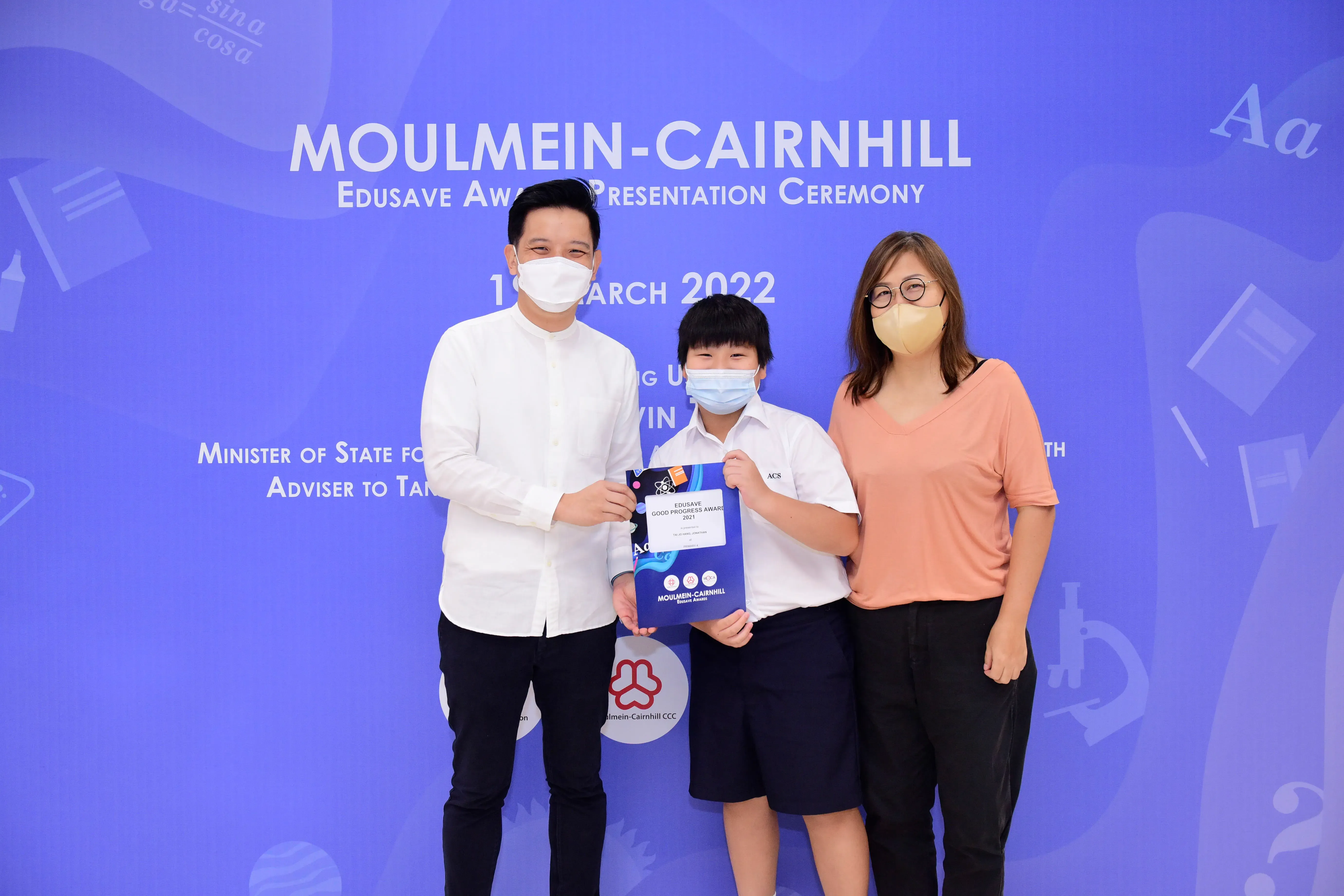 Three masked people pose at the Moulmein-Cairnhill Edusave Award Presentation Ceremony, child holding a certificate.