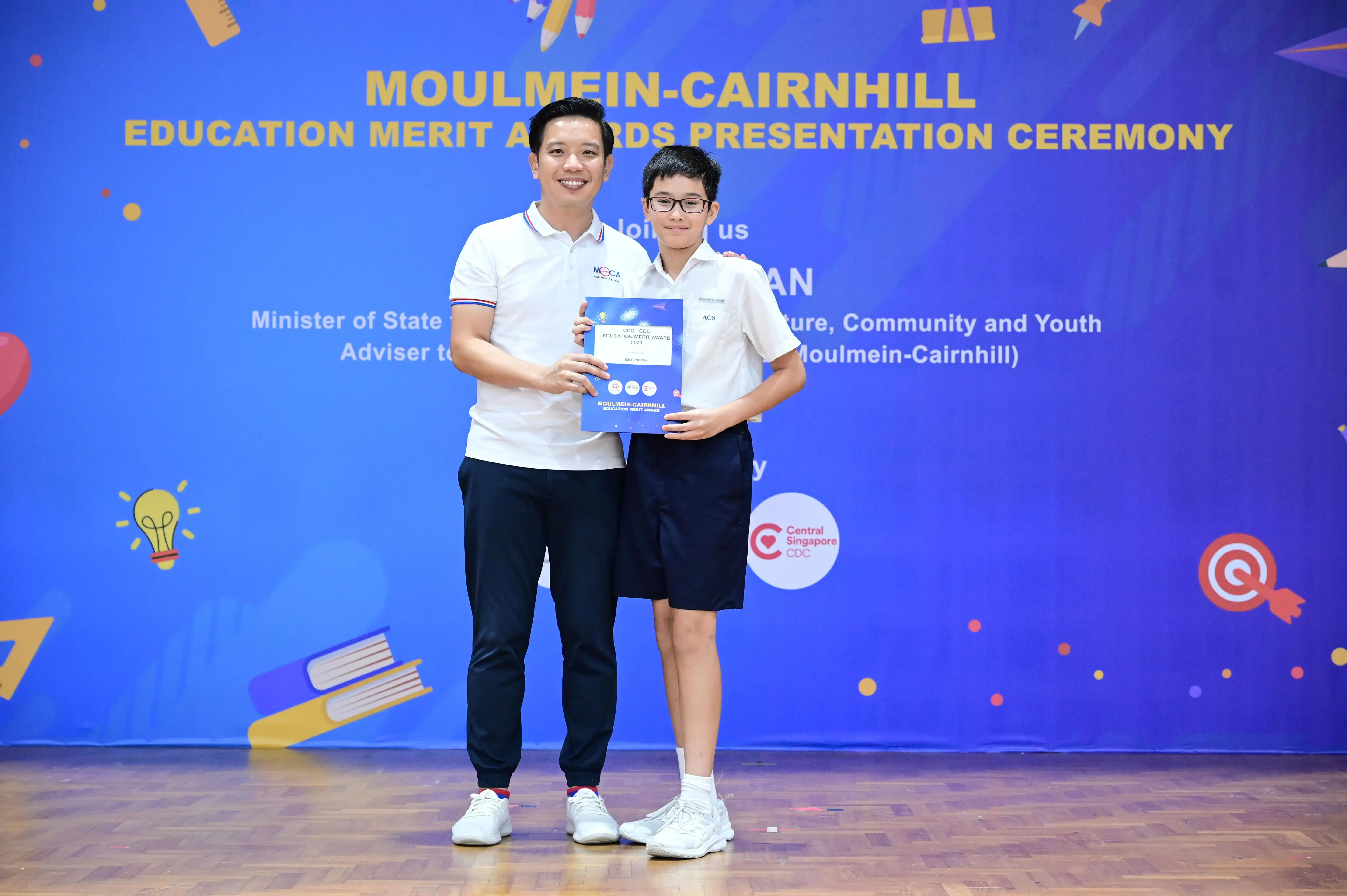 An adult and a young student hold an award certificate at the Moulmein-Cairnhill Education Merit Awards ceremony.