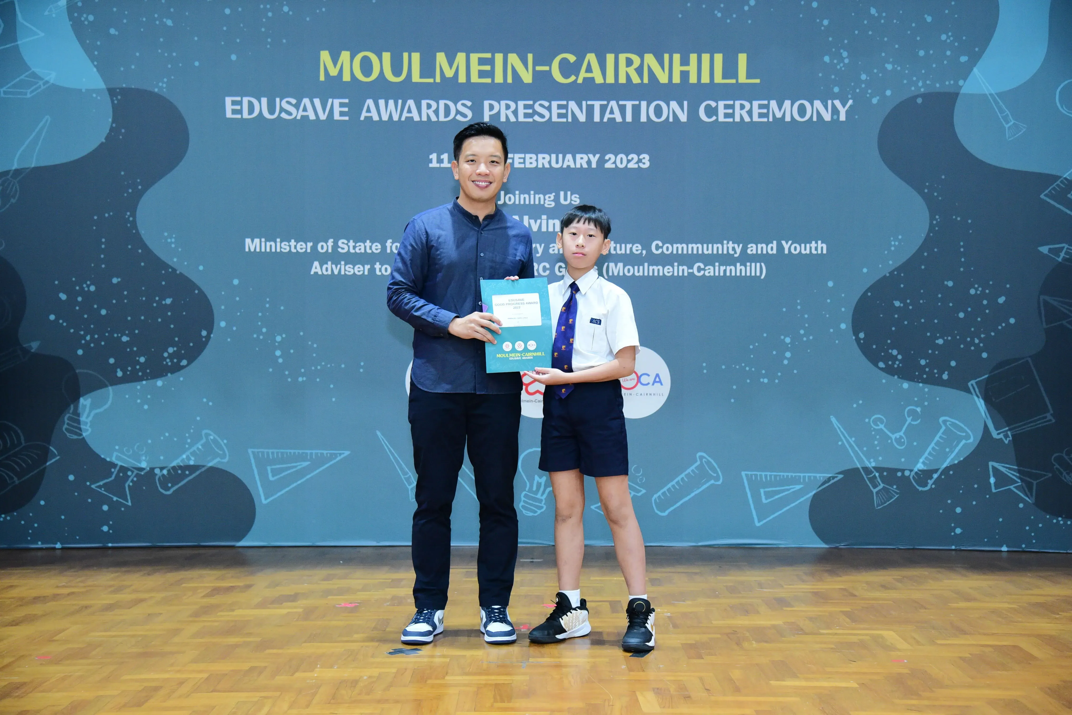 An adult and a young student hold a certificate together at the Moulmein-Cairnhill Edusave Awards Ceremony, February 2023.