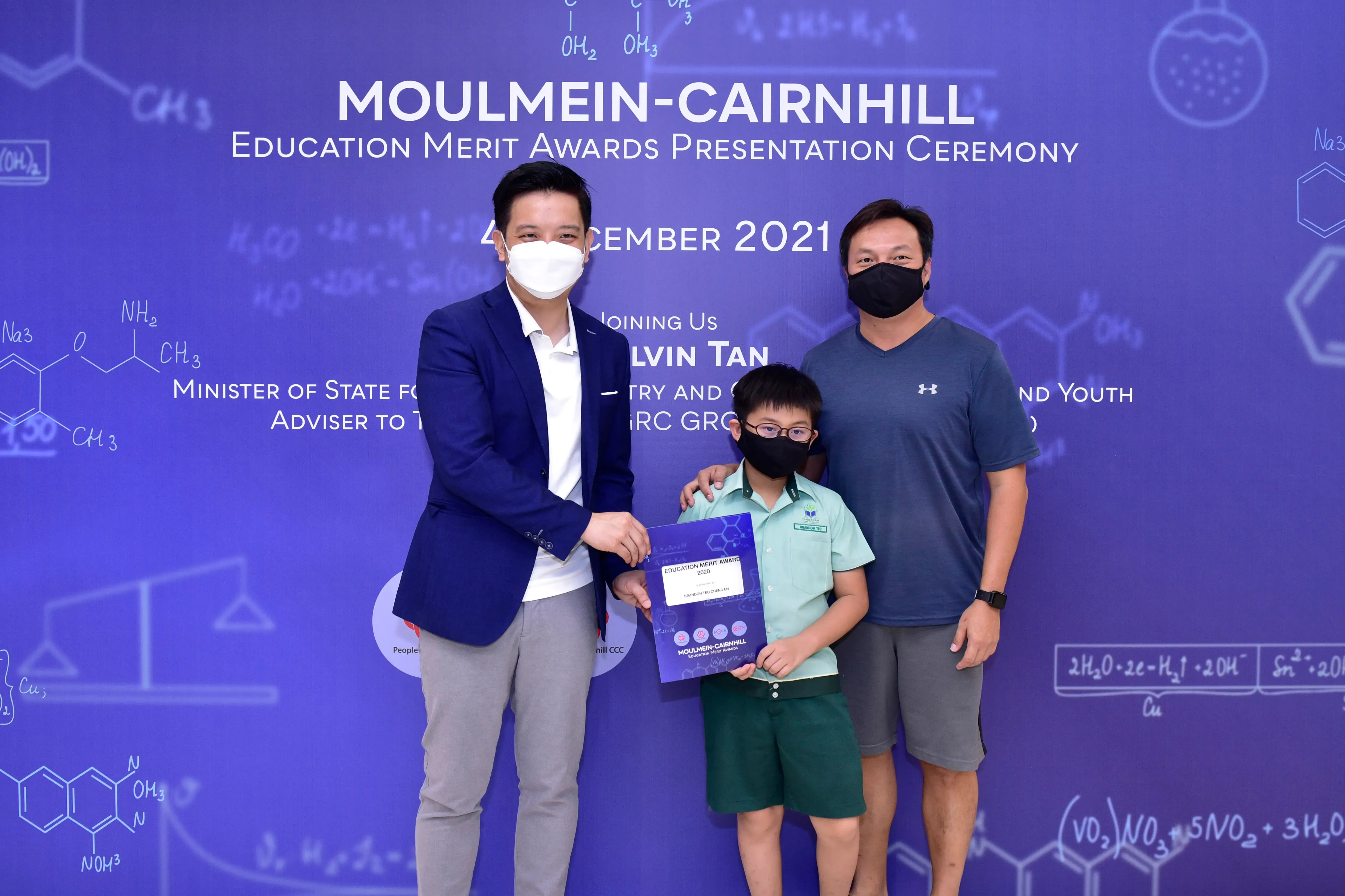 Two masked adults and a masked child hold an award certificate at the Moulmein-Cairnhill Education Merit Awards 2021.
