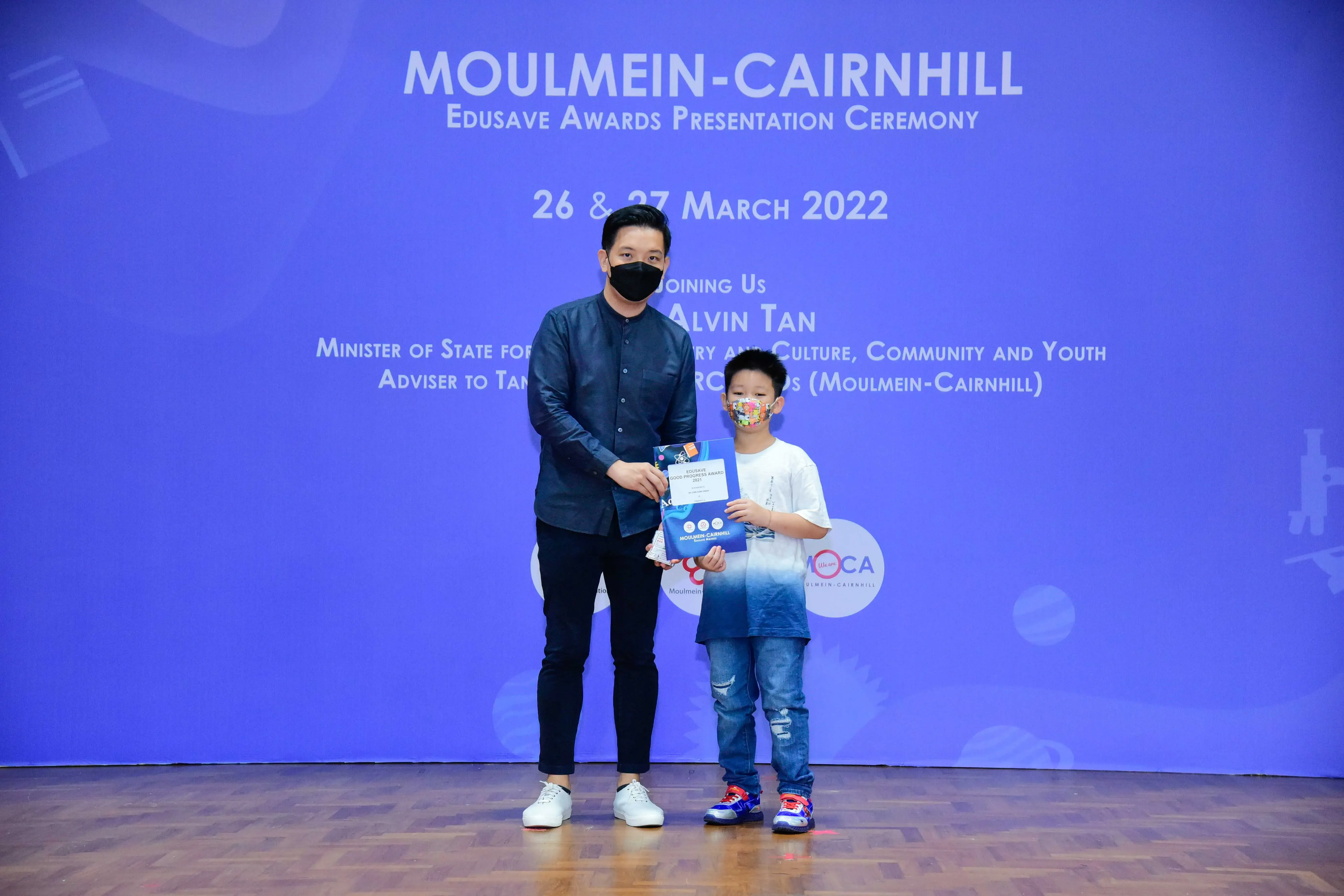 An adult and a young boy wearing masks hold an award certificate at the Moulmein-Cairnhill Edusave Awards Ceremony 2022.
