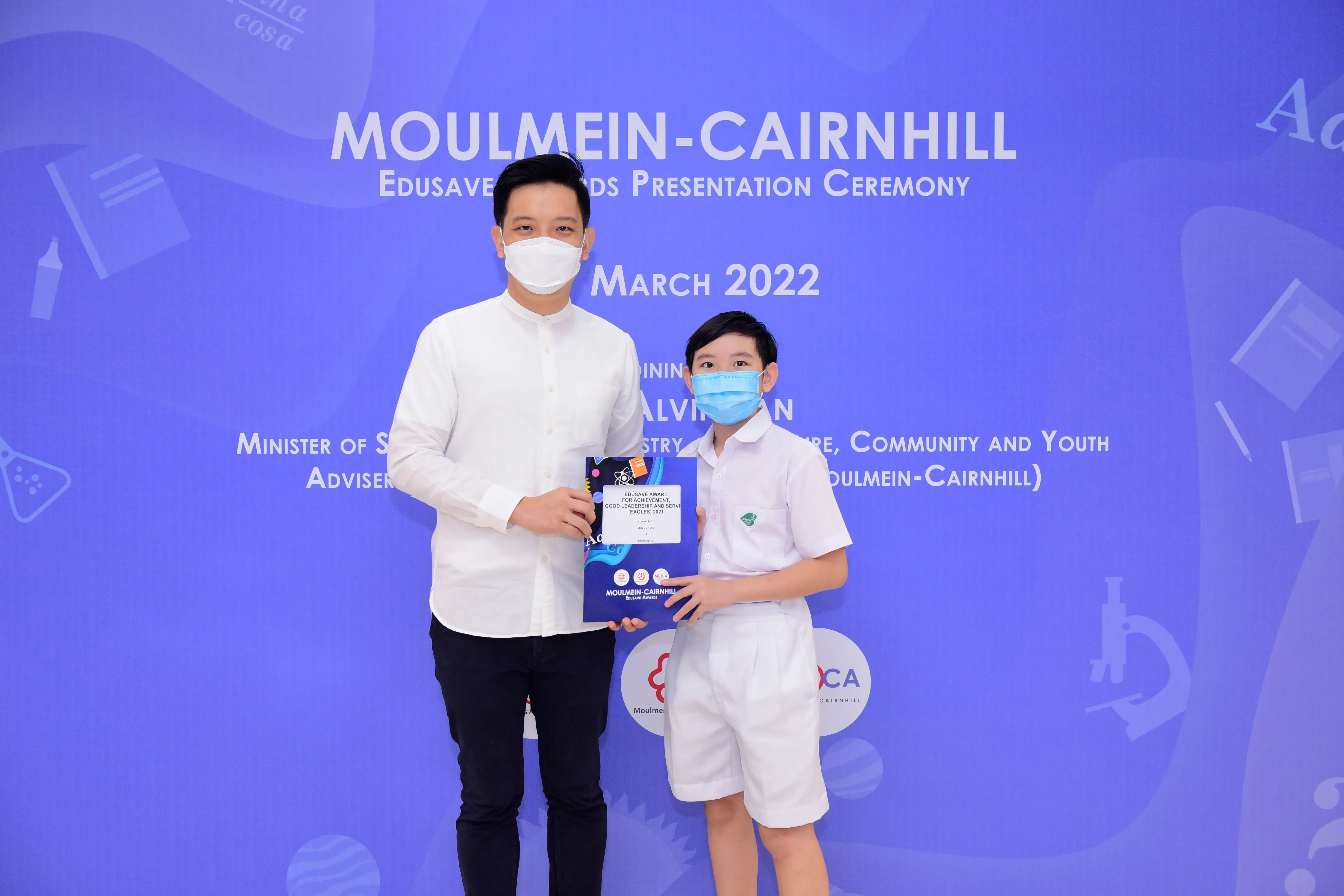 An adult and a young boy wearing masks hold an award at the Moulmein-Cairnhill Edusave Ceremony, March 2022.