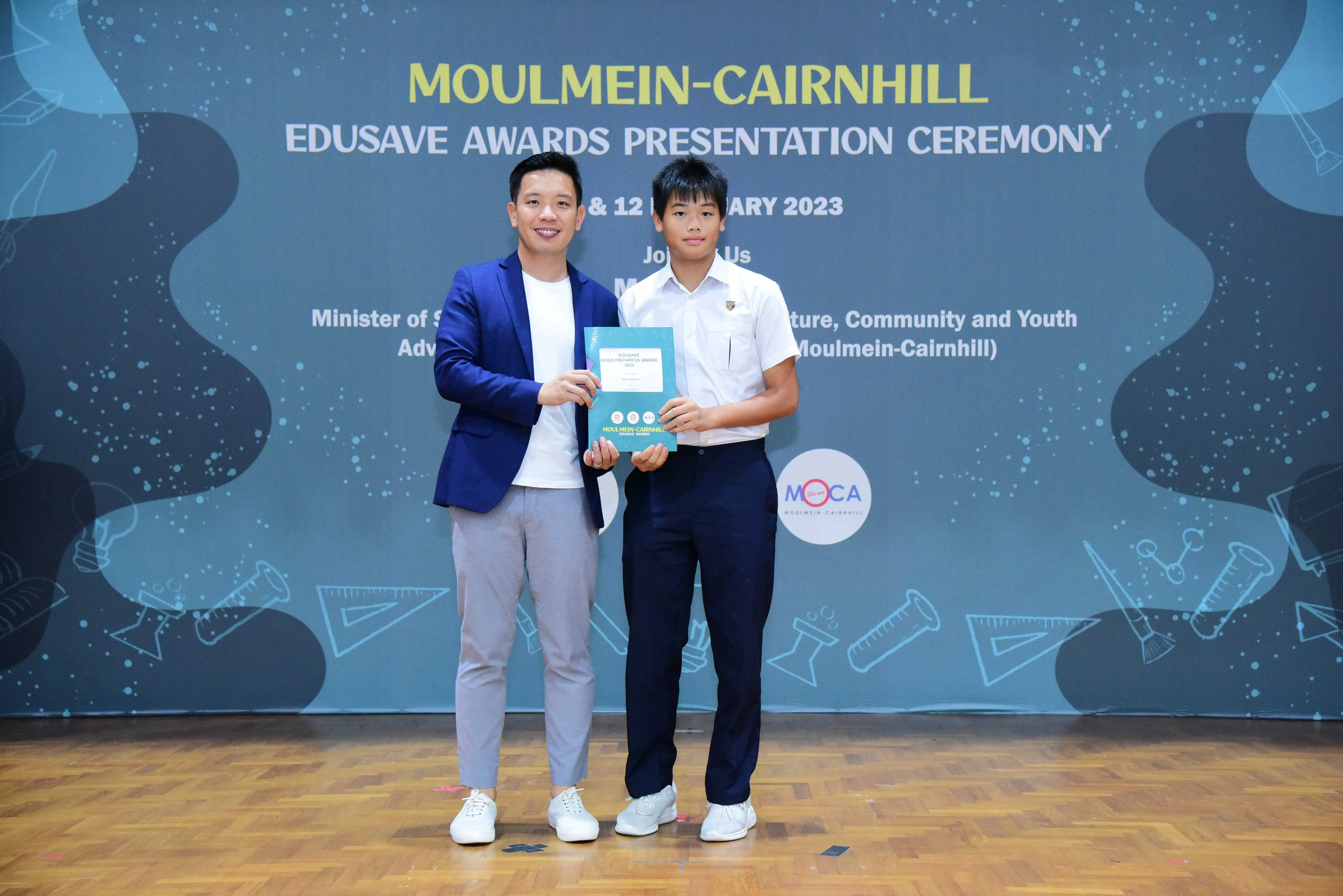 Two males hold a certificate together on stage at the Moulmein-Cairnhill Edusave Awards Presentation Ceremony 2023.