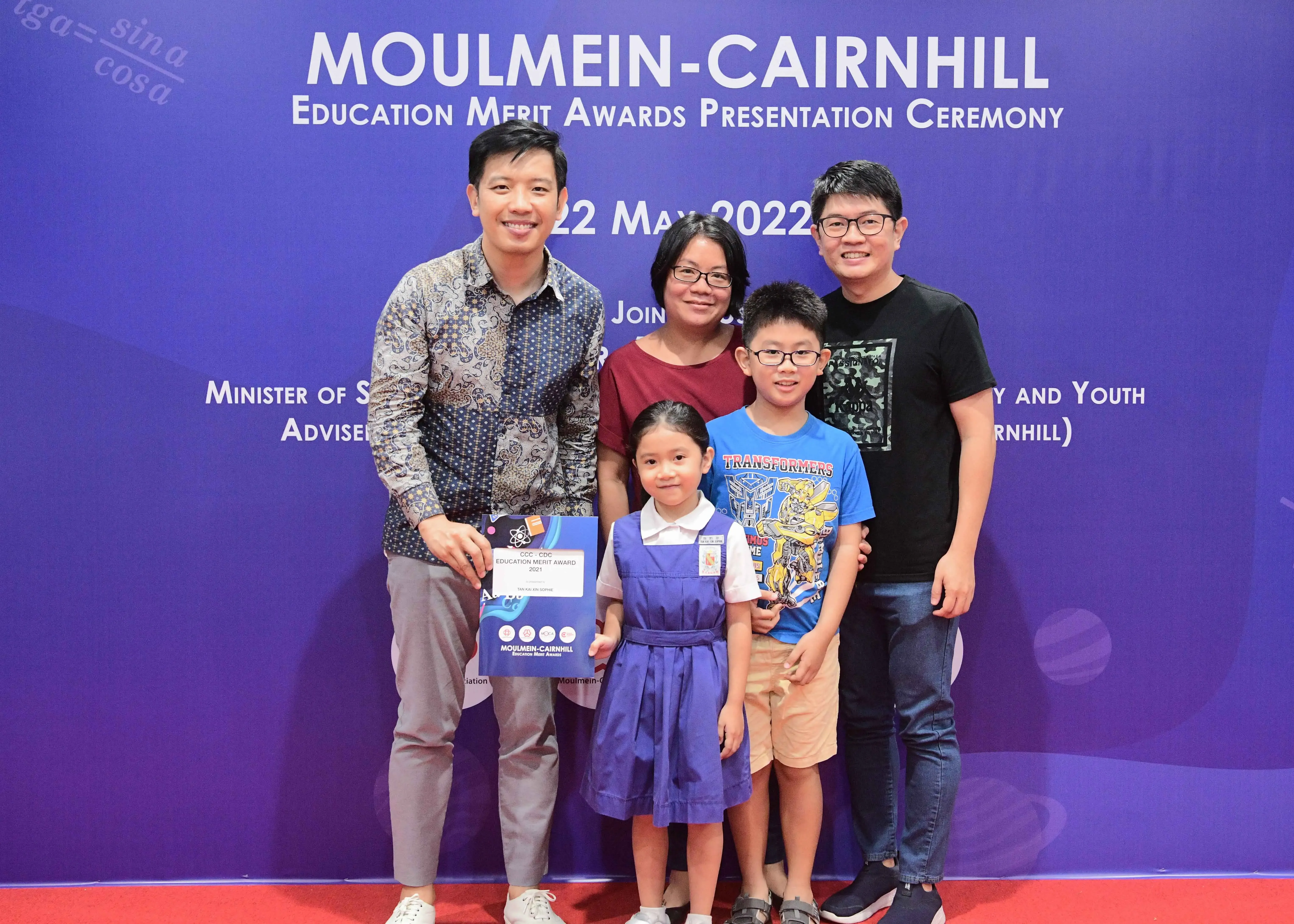 Five people pose in front of a Moulmein-Cairnhill Education Merit Awards banner dated 22 May 2022.
