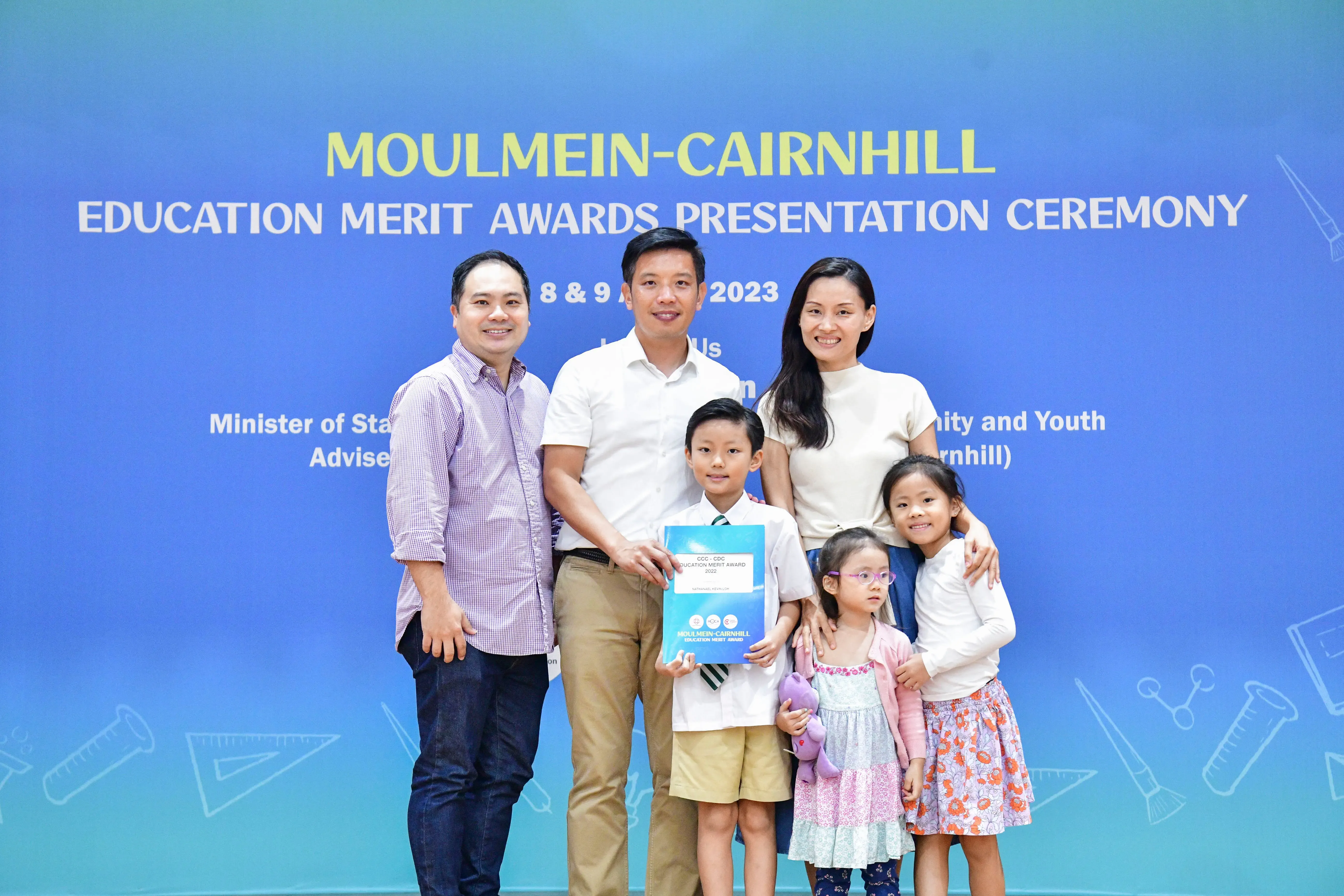 Two adults and three children pose before the Moulmein-Cairnhill Education Merit Awards Ceremony 2023 banner.