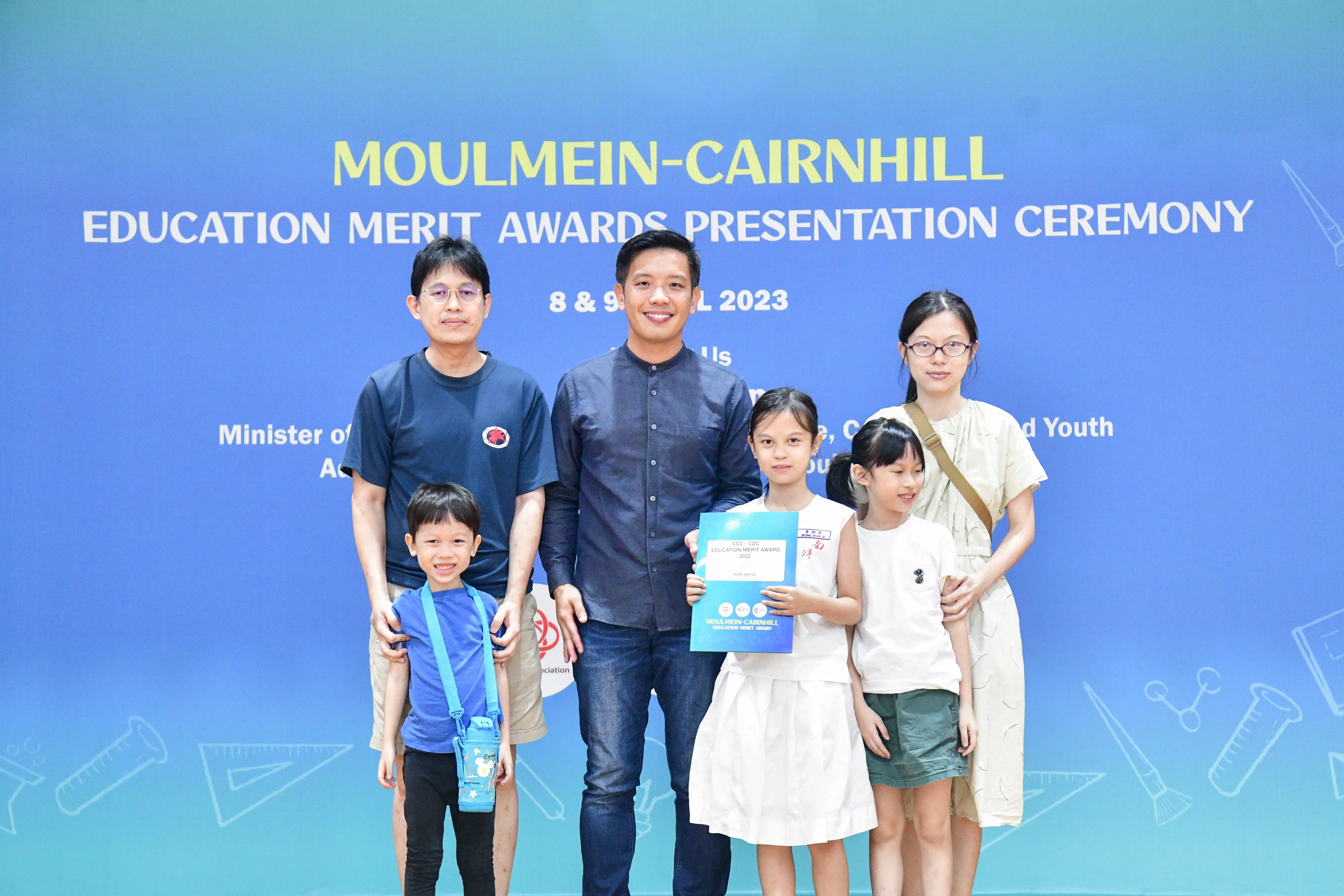 Five people pose in front of a blue Moulmein-Cairnhill Education Merit Awards Presentation Ceremony banner in 2023.