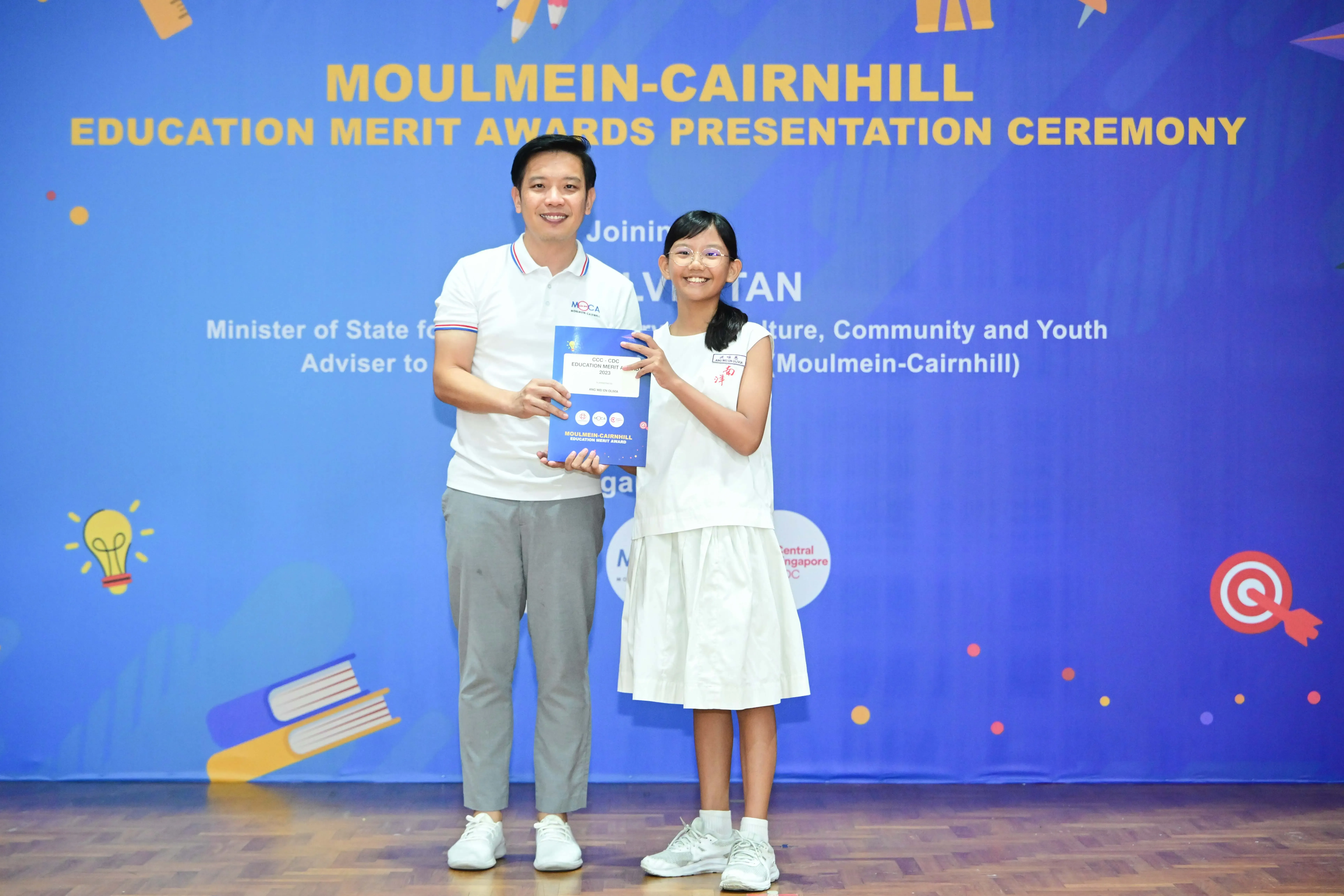 A man and a young girl hold a certificate together at the Moulmein-Cairnhill Education Merit Awards Presentation Ceremony.