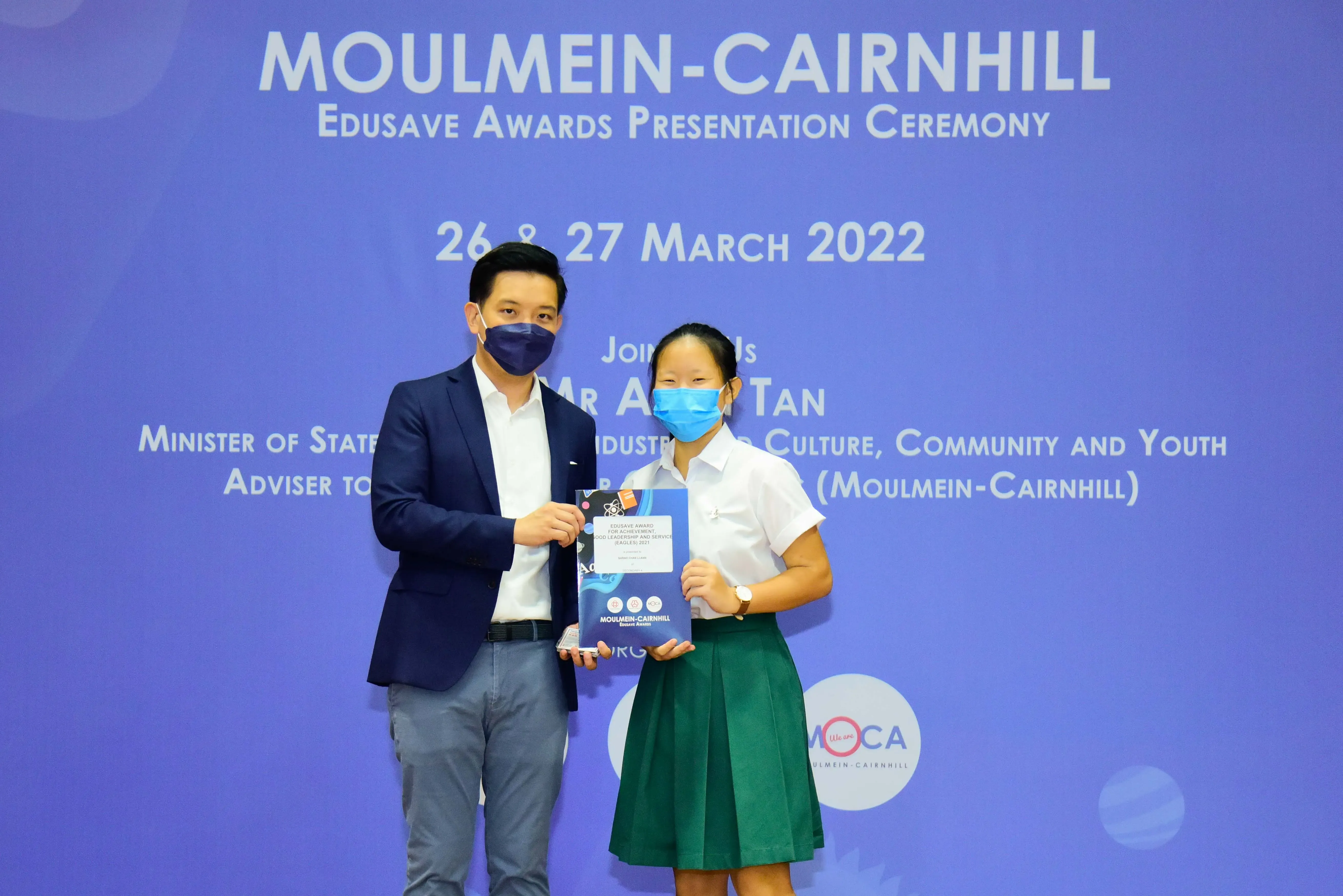 Two masked people hold an award booklet at the Moulmein-Cairnhill Edusave Awards Presentation Ceremony, March 2022.