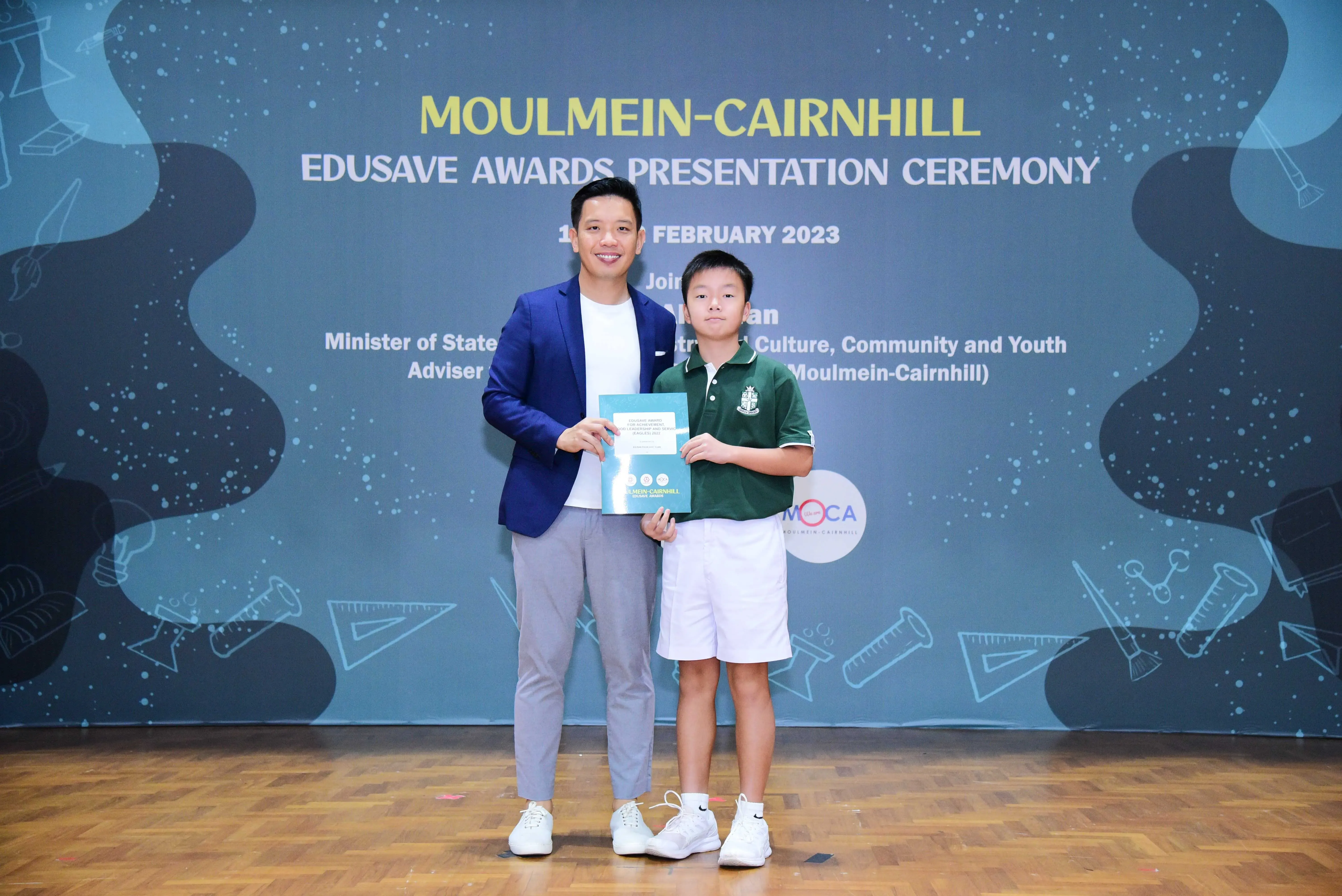 An adult in a blue blazer and a young boy in a green school uniform hold a certificate at the Moulmein-Cairnhill Edusave Awards 2023.