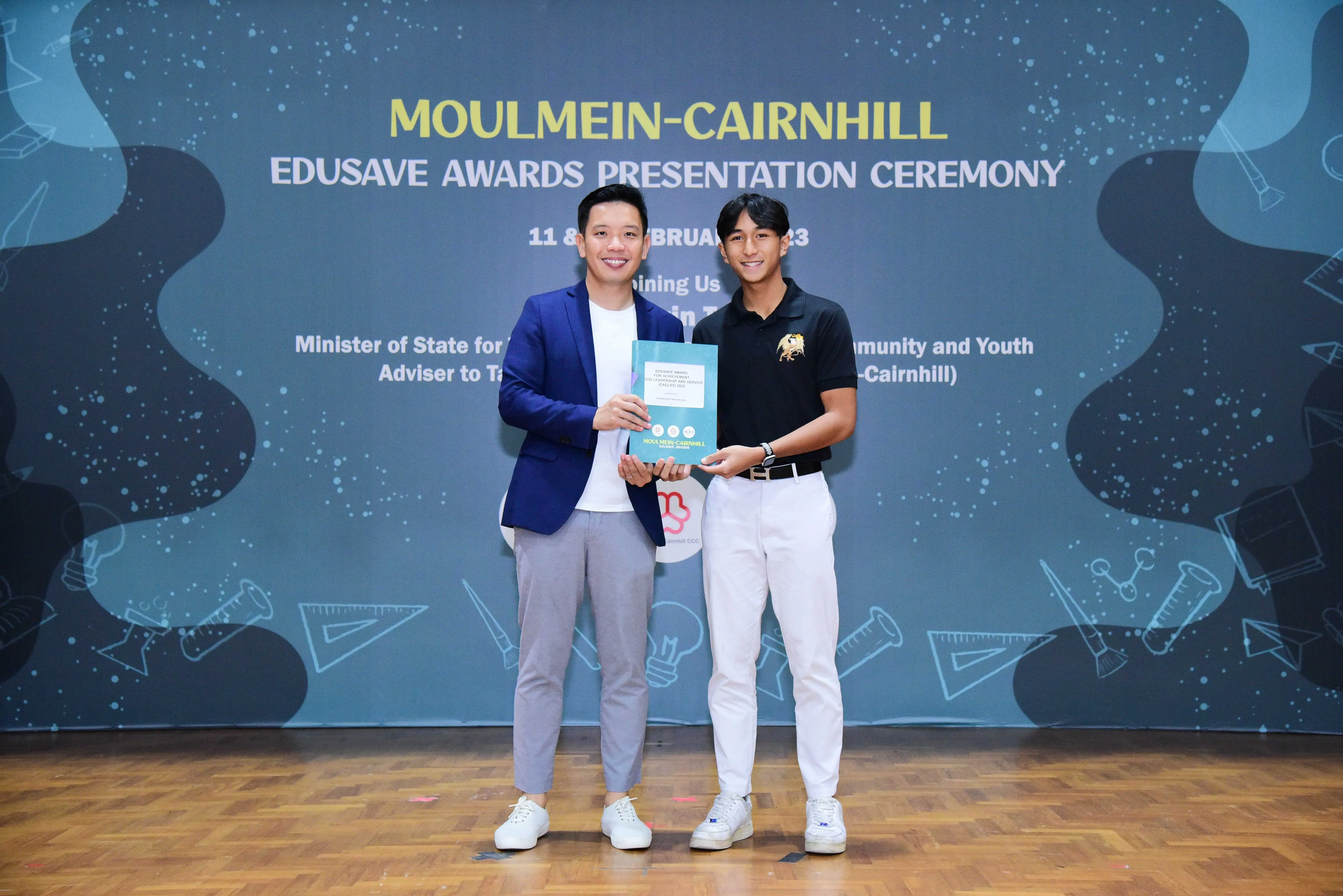 Two men smile holding a certificate at the Moulmein-Cairnhill Edusave Awards Presentation Ceremony backdrop.