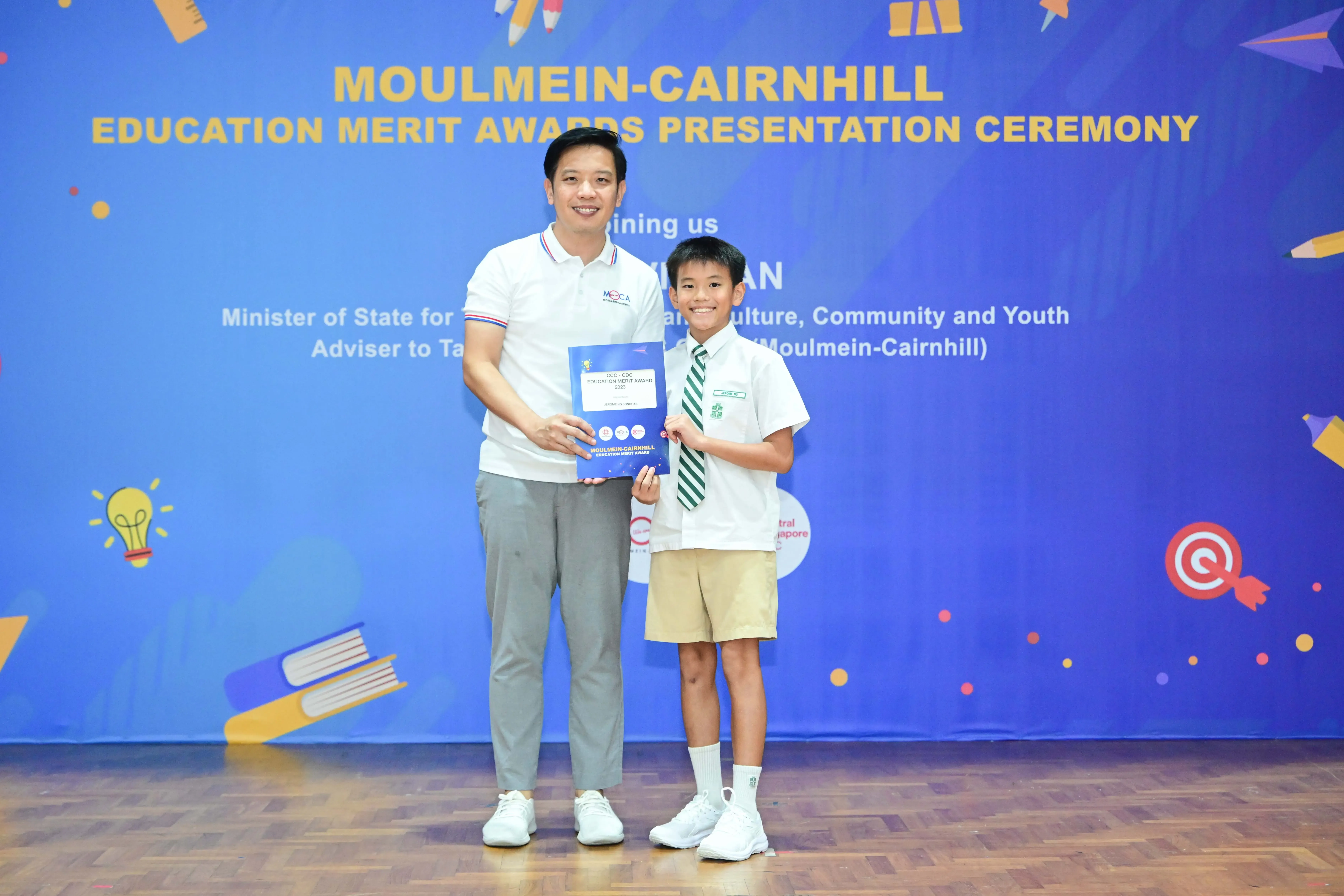 An adult and a young boy in school uniform hold an award certificate at the Moulmein-Cairnhill Education Merit Awards ceremony.